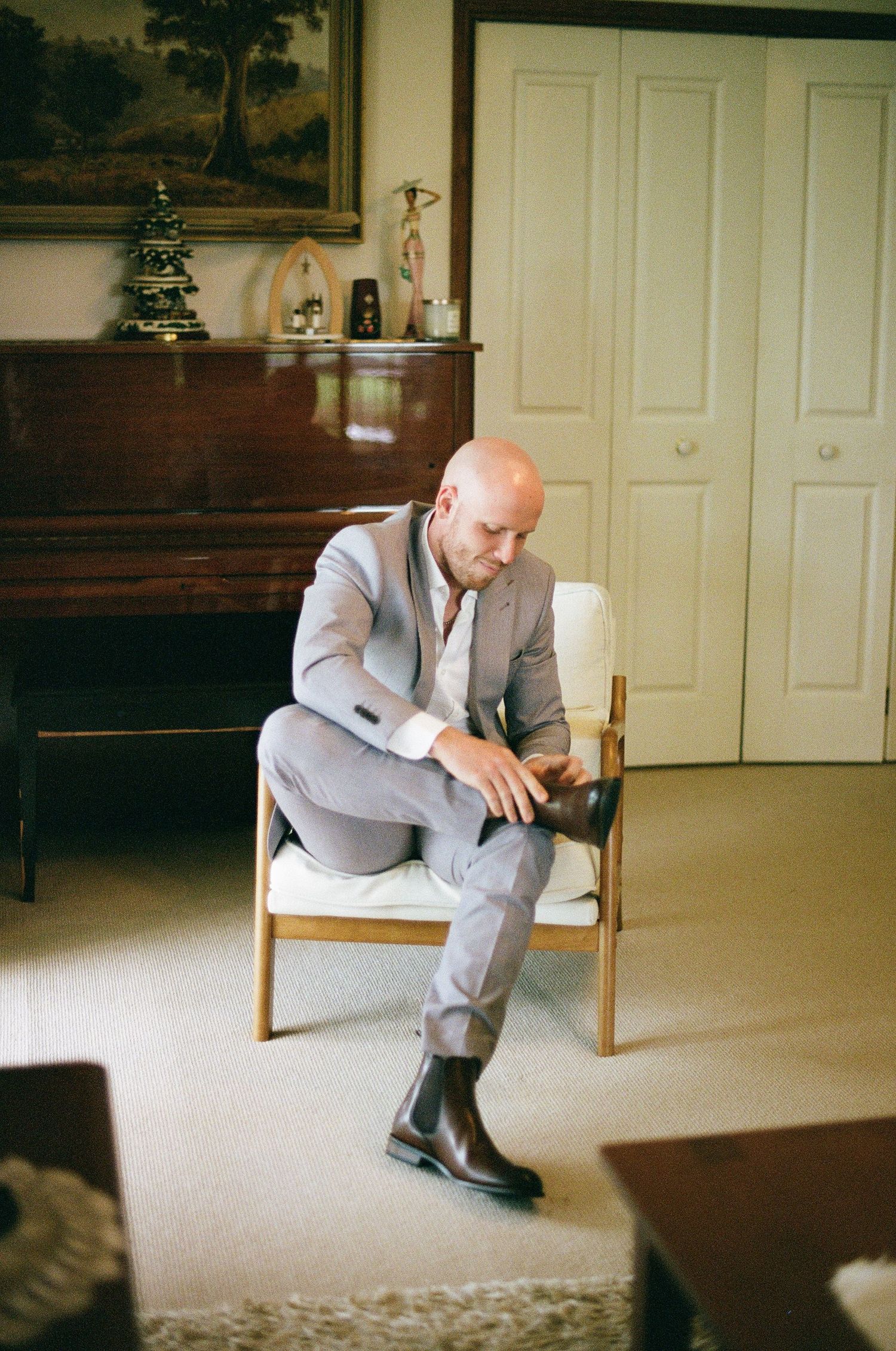 Bald groom in gray suit sitting in wooden chair putting on brown Chelsea boots in elegant room.