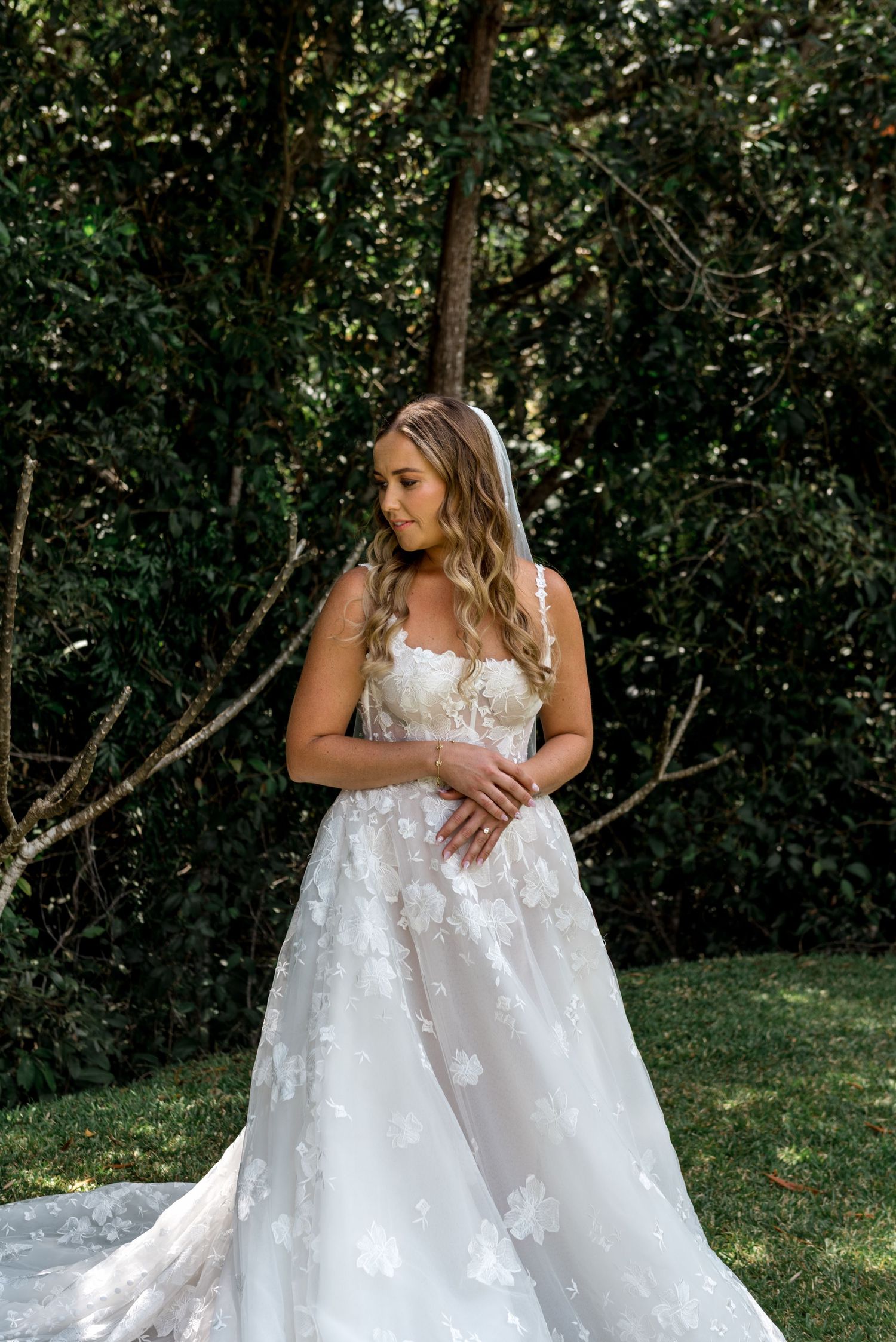 Elegant bride in strapless floral lace wedding dress with cathedral train posing in a garden setting.