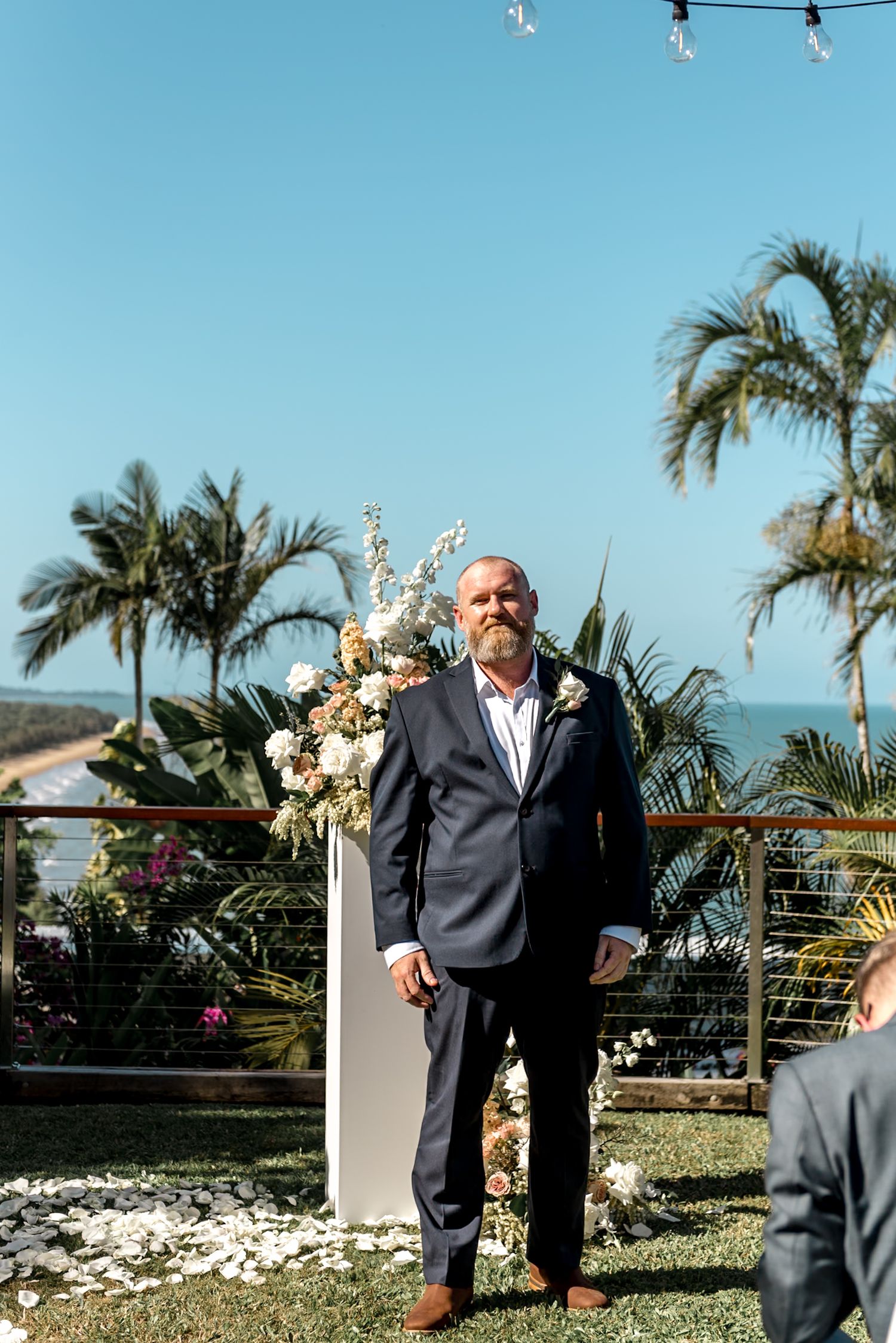 Bearded groom in navy suit stands at outdoor wedding altar with floral arrangements and ocean view.