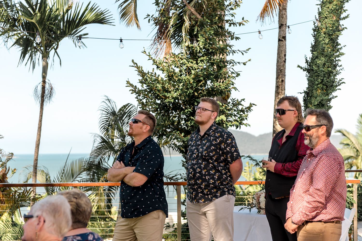 Four male wedding guests watch outdoor ceremony near ocean with palm trees and string lights overhead.