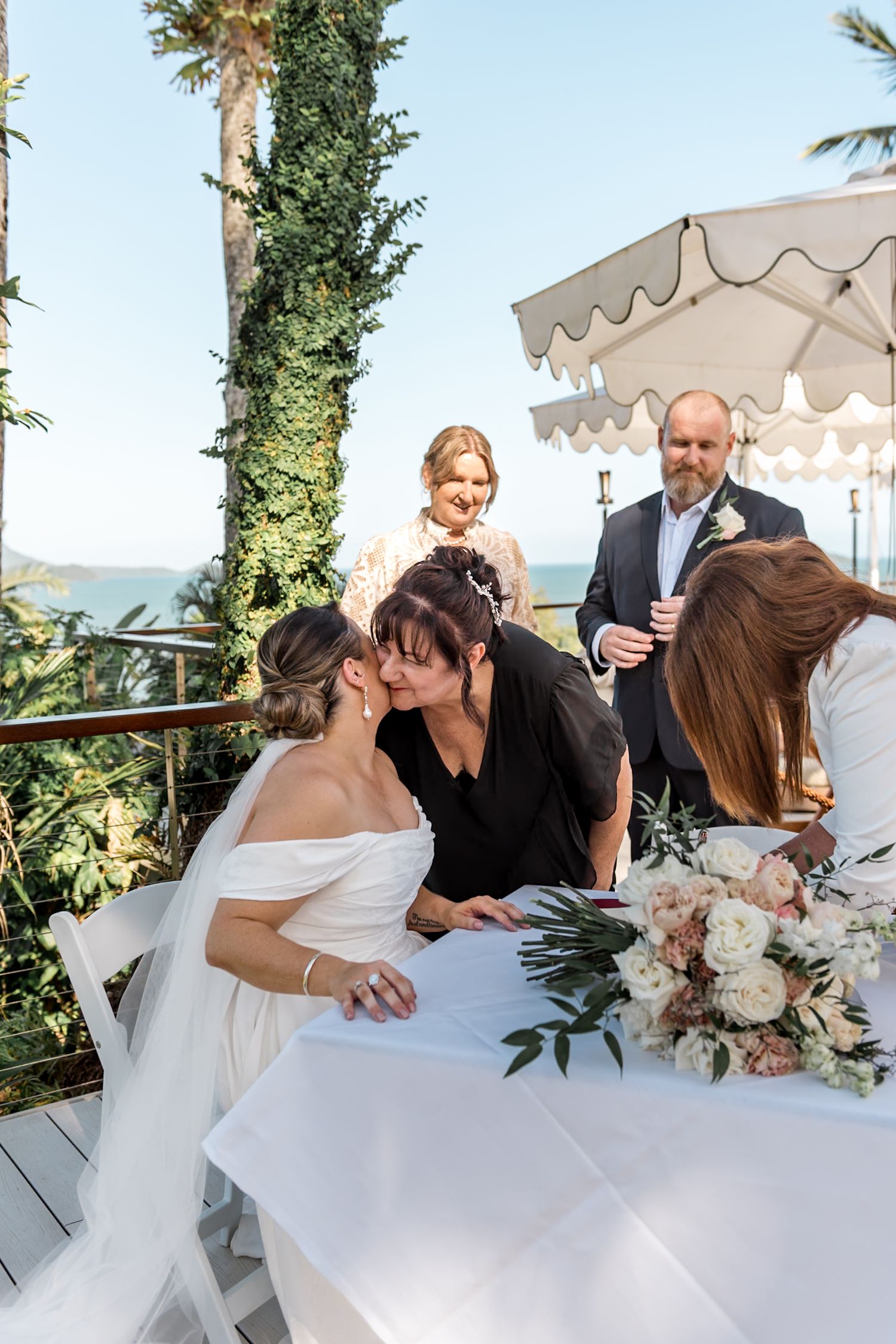 Bride receiving congratulatory kiss while signing marriage certificate at tropical outdoor venue.