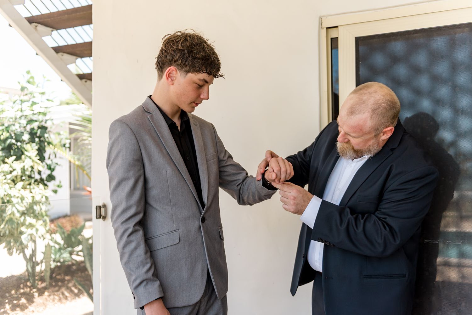 Older man helping young groom in gray suit fasten his cufflinks outdoors.