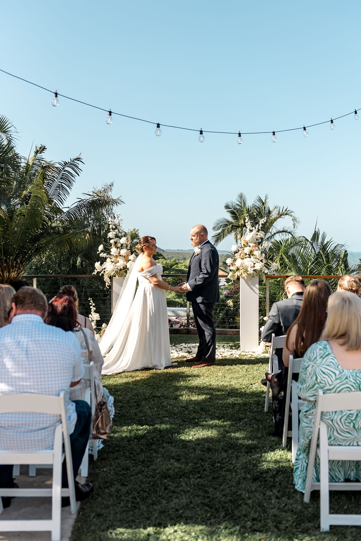 Bride and groom stand at floral altar during outdoor wedding ceremony with string lights and tropical palms.