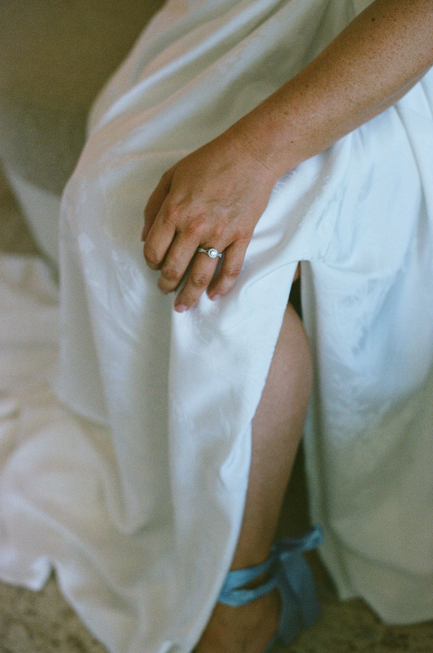Close-up of bride's hand with diamond engagement ring resting on white wedding dress with blue garter visible.