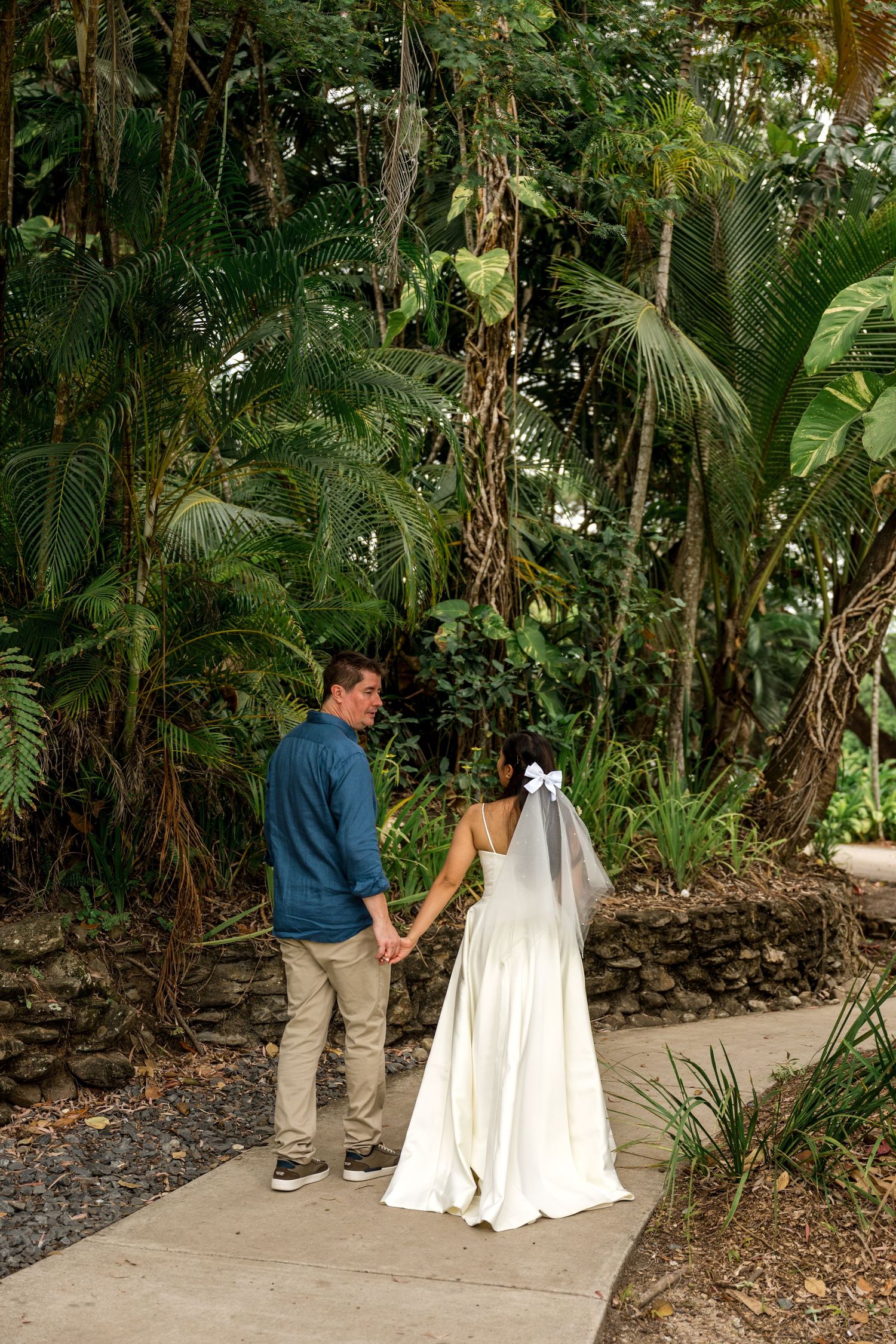 Bride and groom hold hands on a garden path surrounded by lush tropical greenery at their outdoor wedding at Little Cove, Port Douglas.