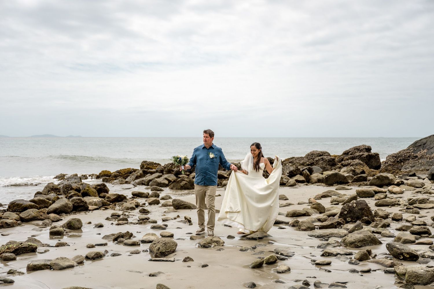 Bride and groom walking hand in hand along a rocky beach on their wedding day at Little Cove, Port Douglas.