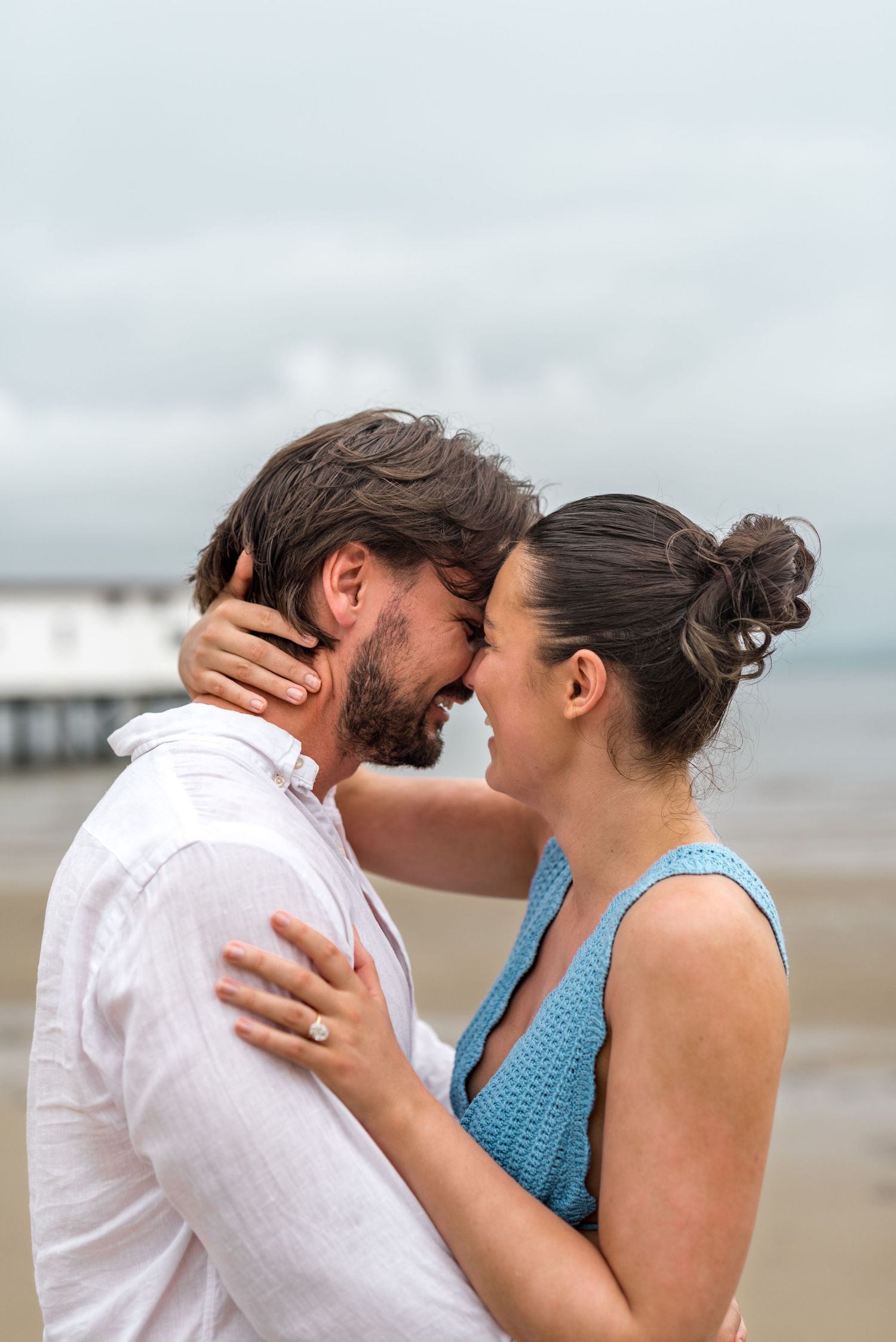 Close-up of a smiling couple touching foreheads on a beach, with a diamond engagement ring visible on her hand.