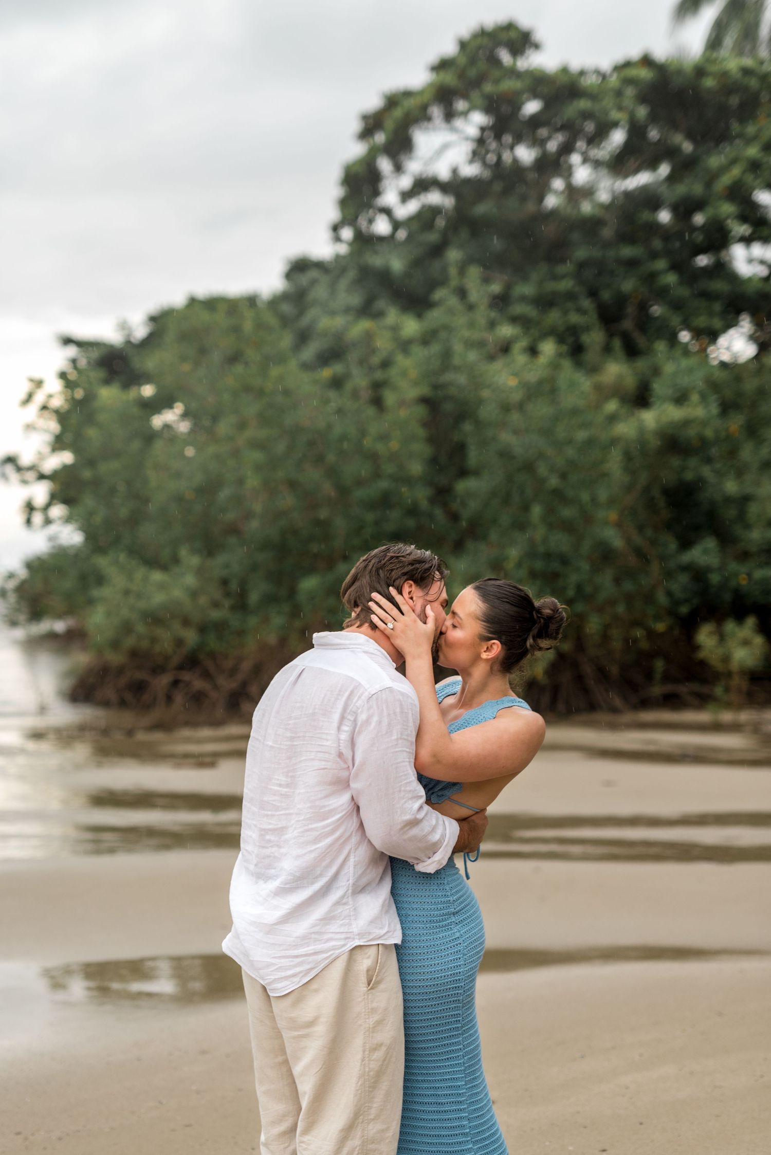 Couple sharing a romantic kiss on a tropical beach with lush green trees in the background.