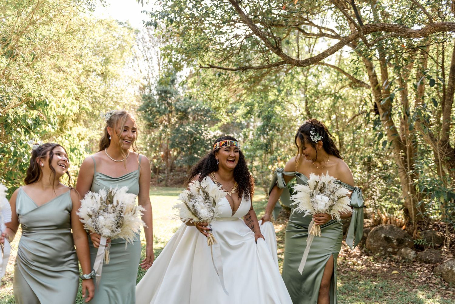 A bride and three bridesmaids in sage green dresses walk and laugh in a sunny garden.