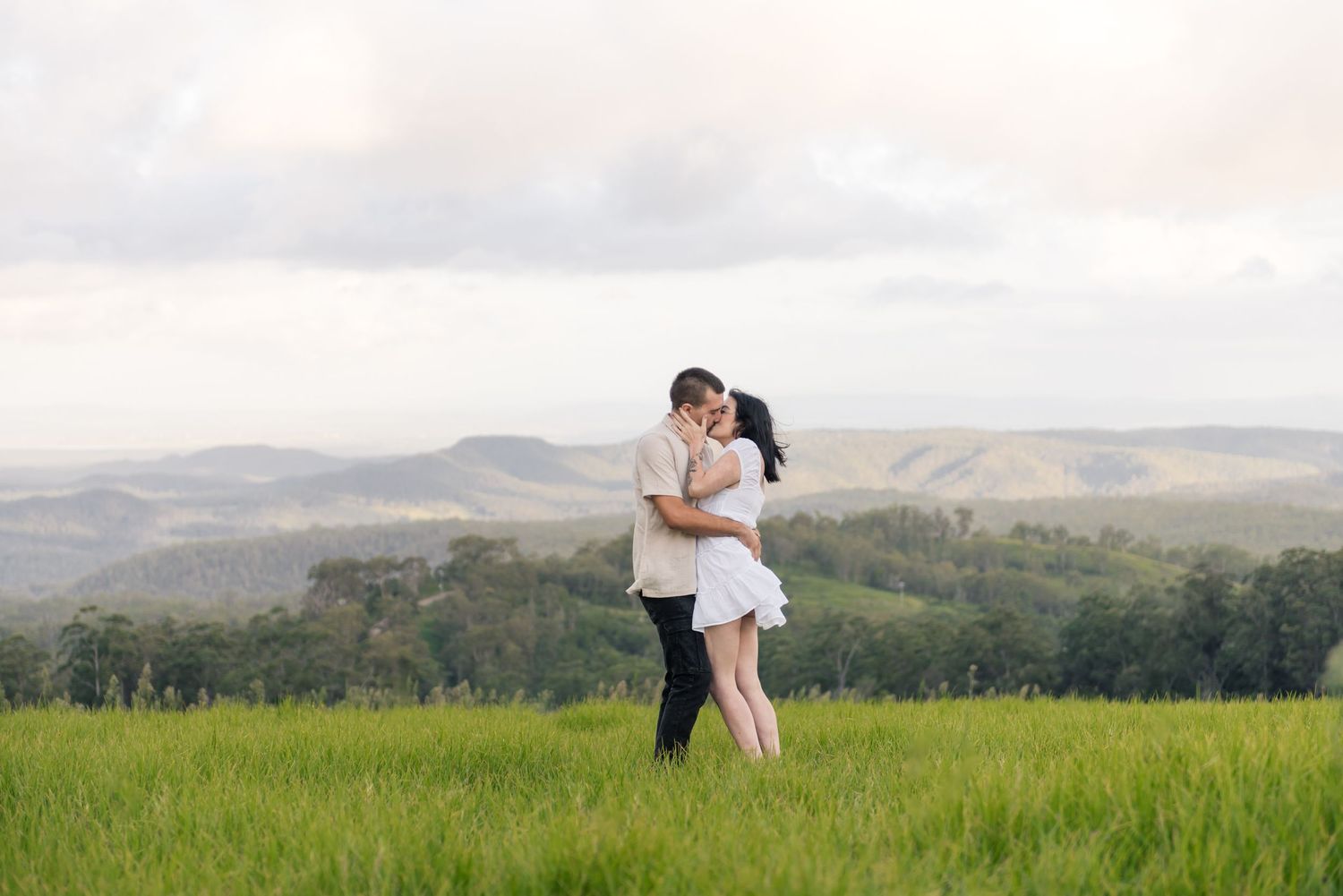 Romantic couple embracing and kissing in a green field with stunning mountain scenery.
