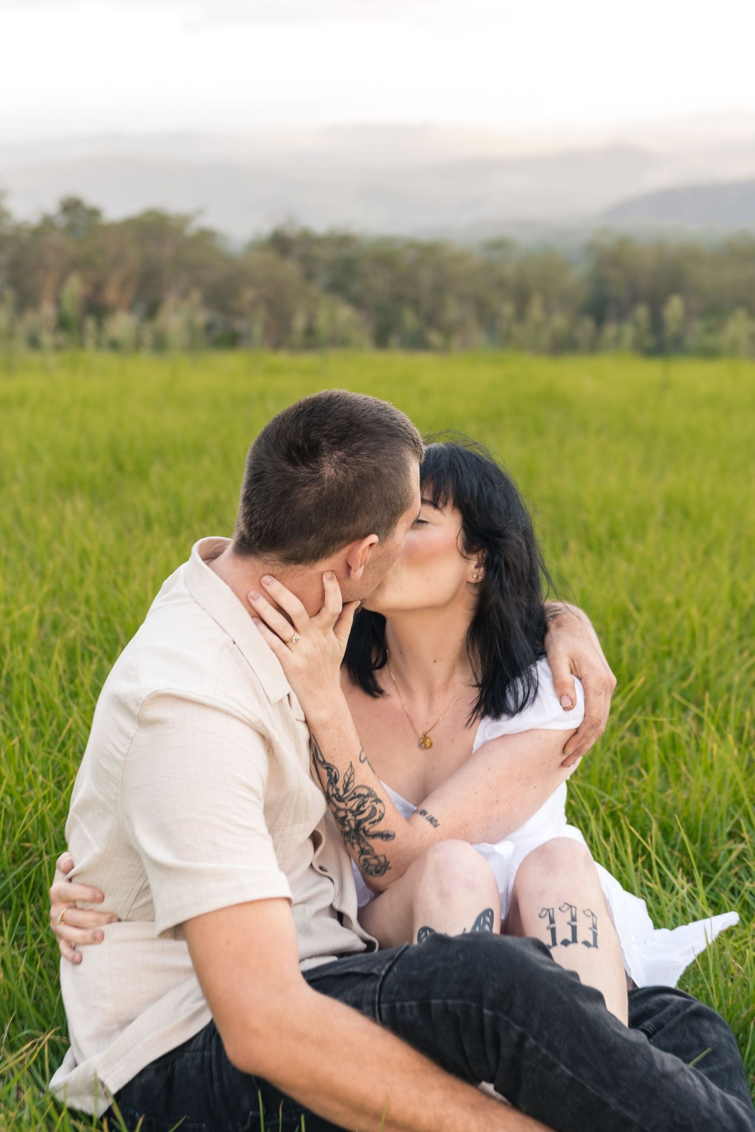 Couple sharing a kiss while sitting in lush green grass field with mountains in background.