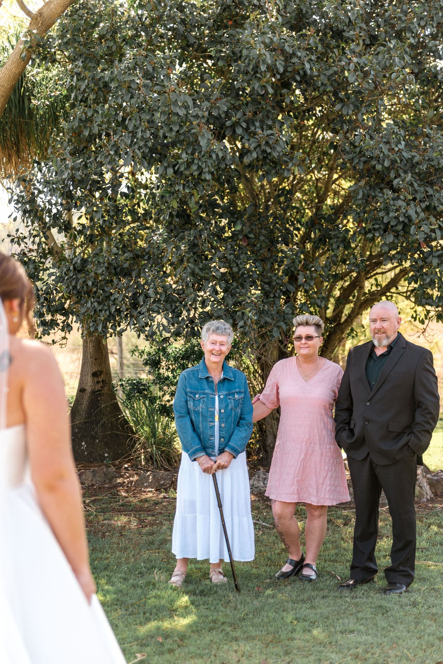 Bride in white gown shares emotional first look moment with elderly woman and couple outdoors.