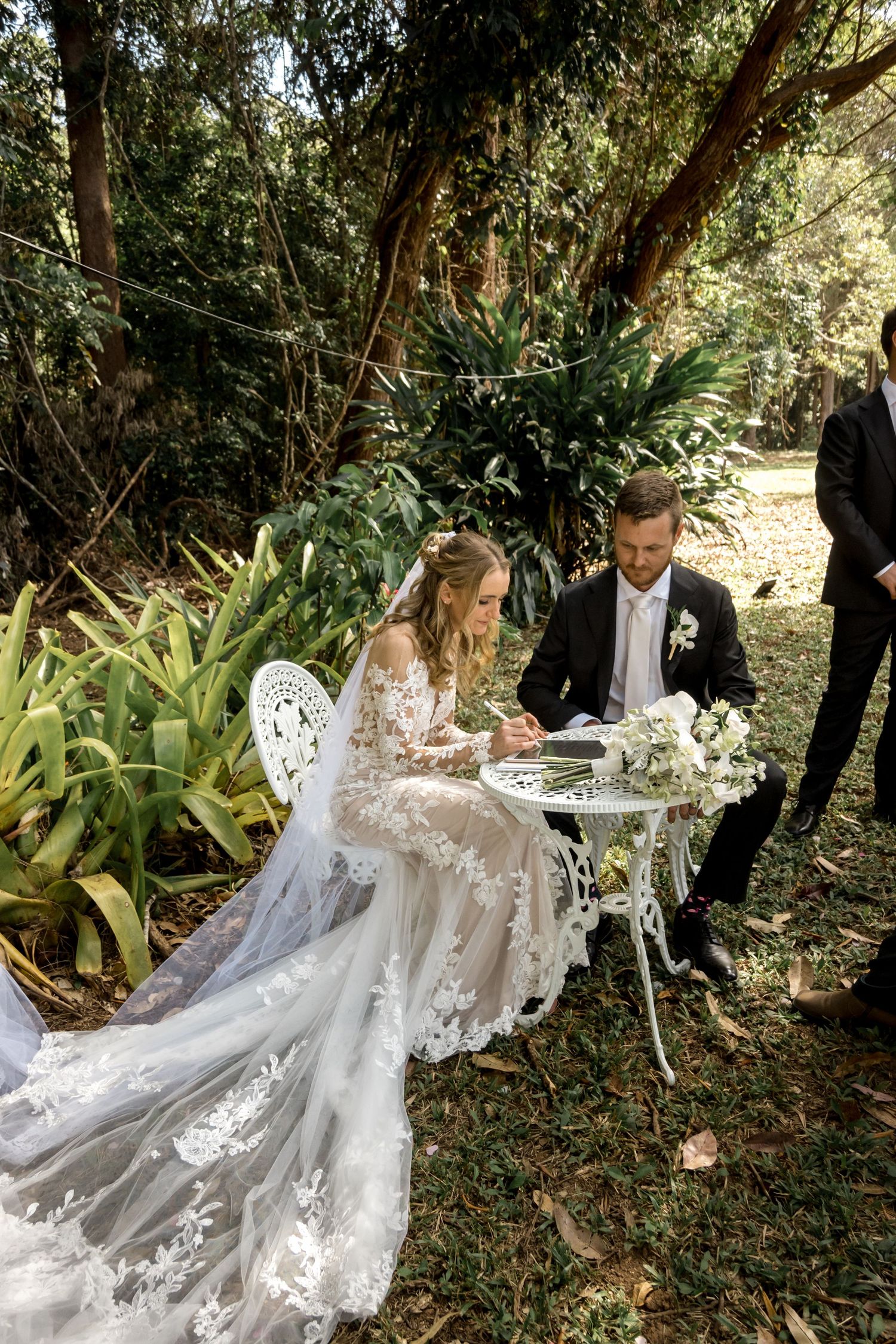 Bride and groom sign wedding certificate at ornate white table in lush garden setting.