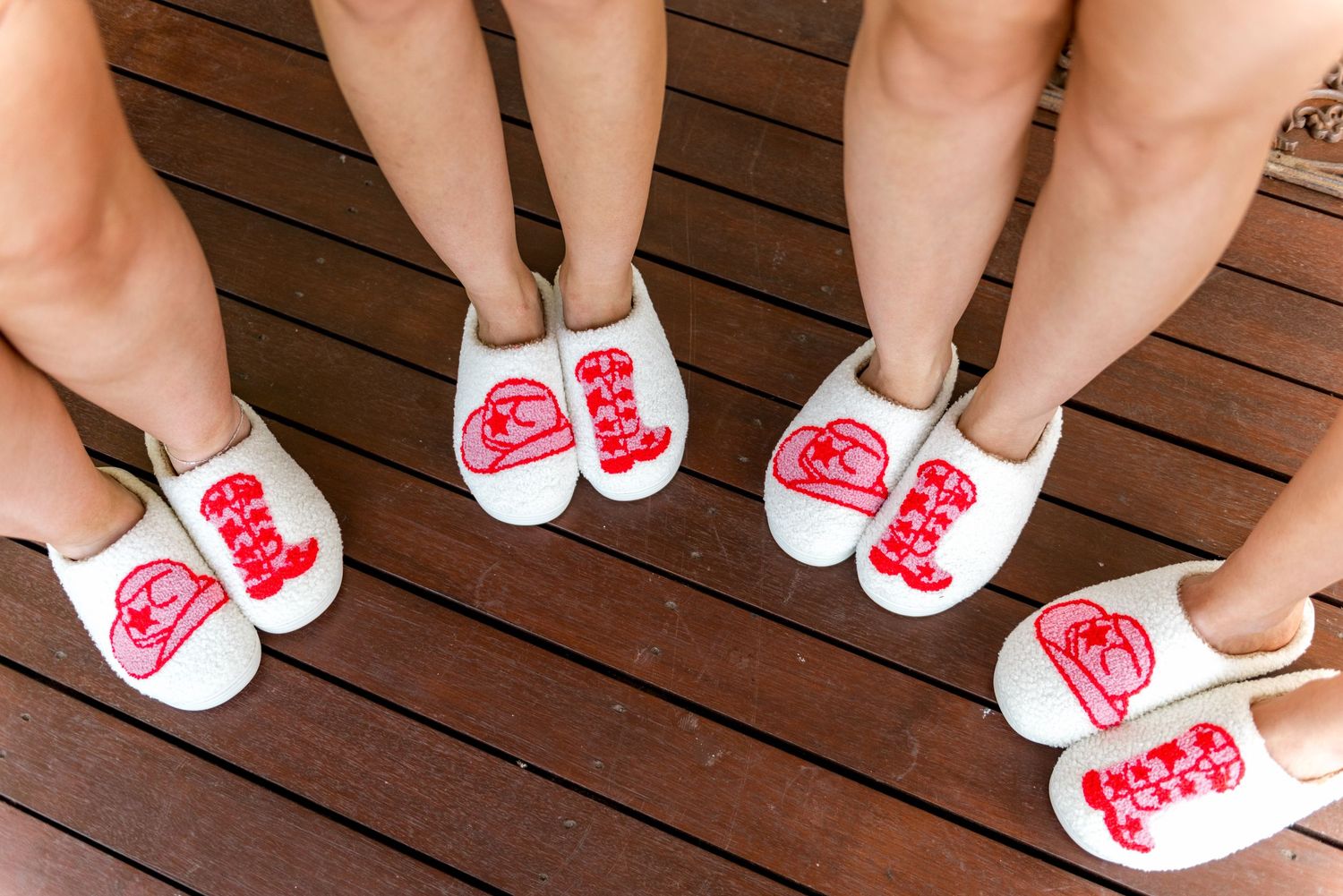 Four pairs of feet wearing personalised white and red embroidered wedding slippers on timber deck.