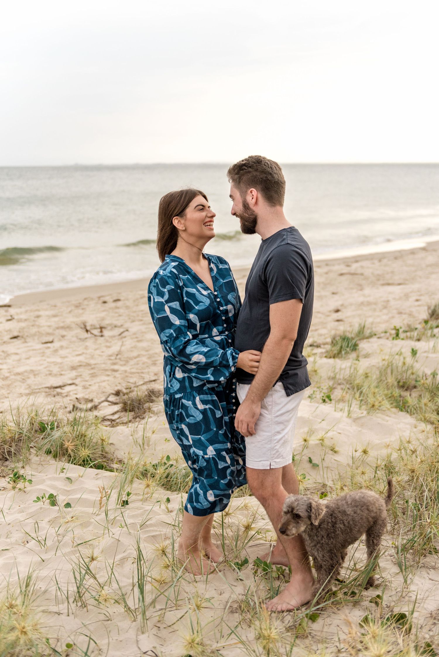 Couple in love standing together on beach dunes with grey dog beside them.