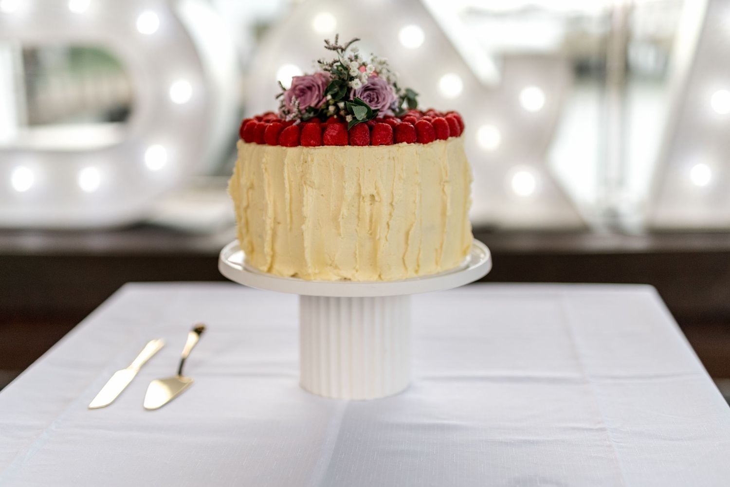 White wedding cake with raspberry topping and floral arrangement displayed on pedestal cake stand.