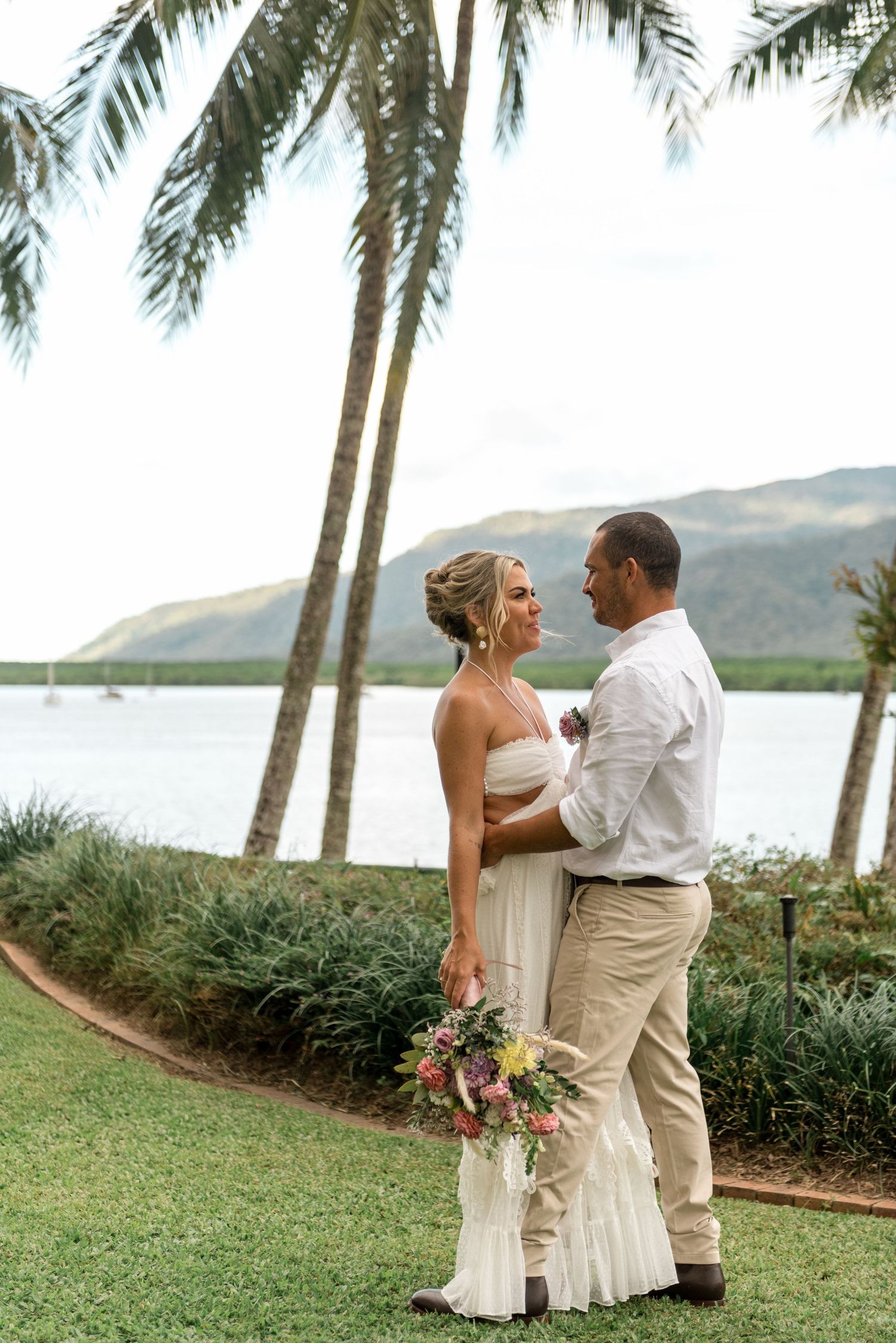 Bride and groom share a romantic moment by a tropical waterway surrounded by lush palm trees.