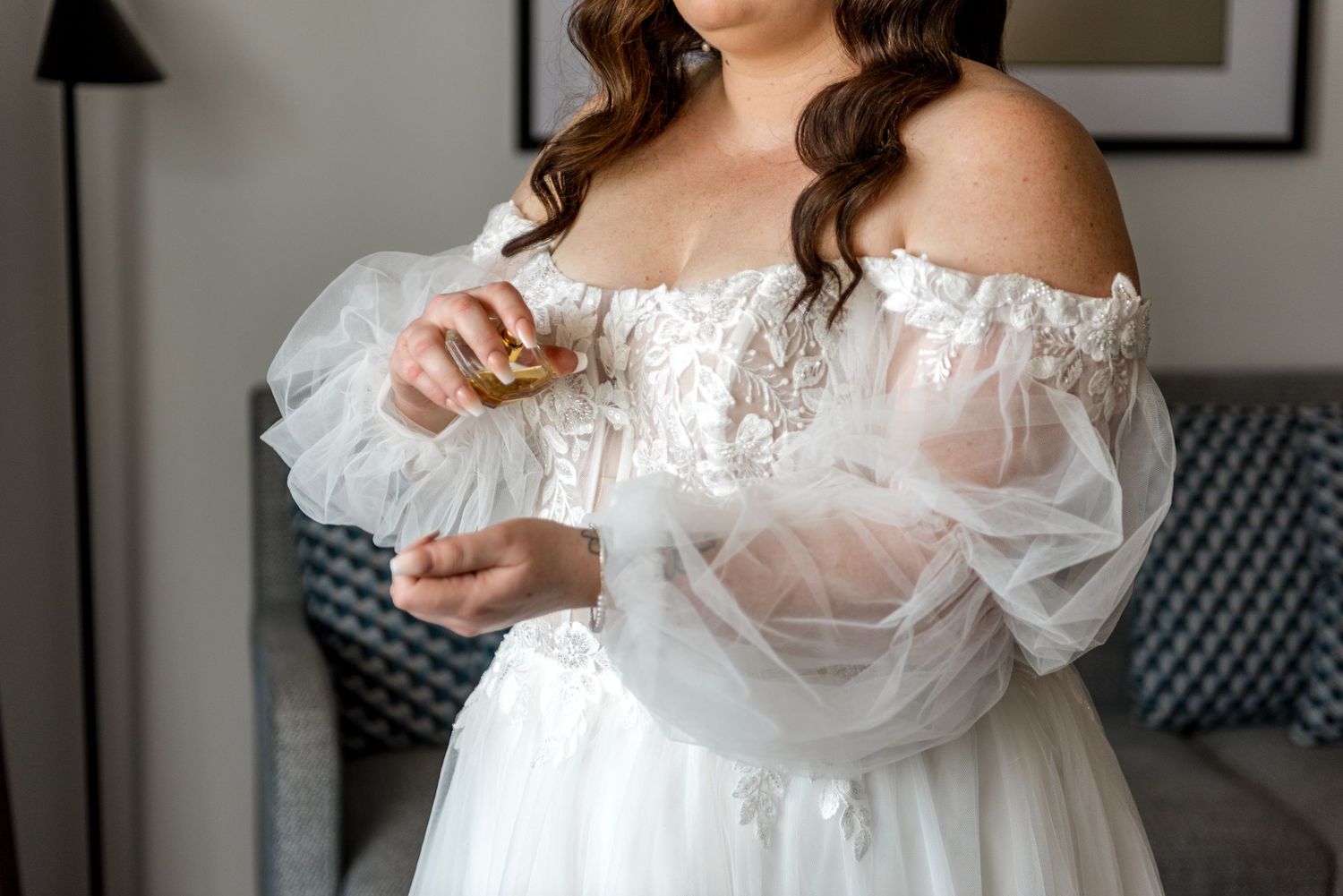 Bride in off-shoulder lace tulle wedding dress applying perfume in hotel room.