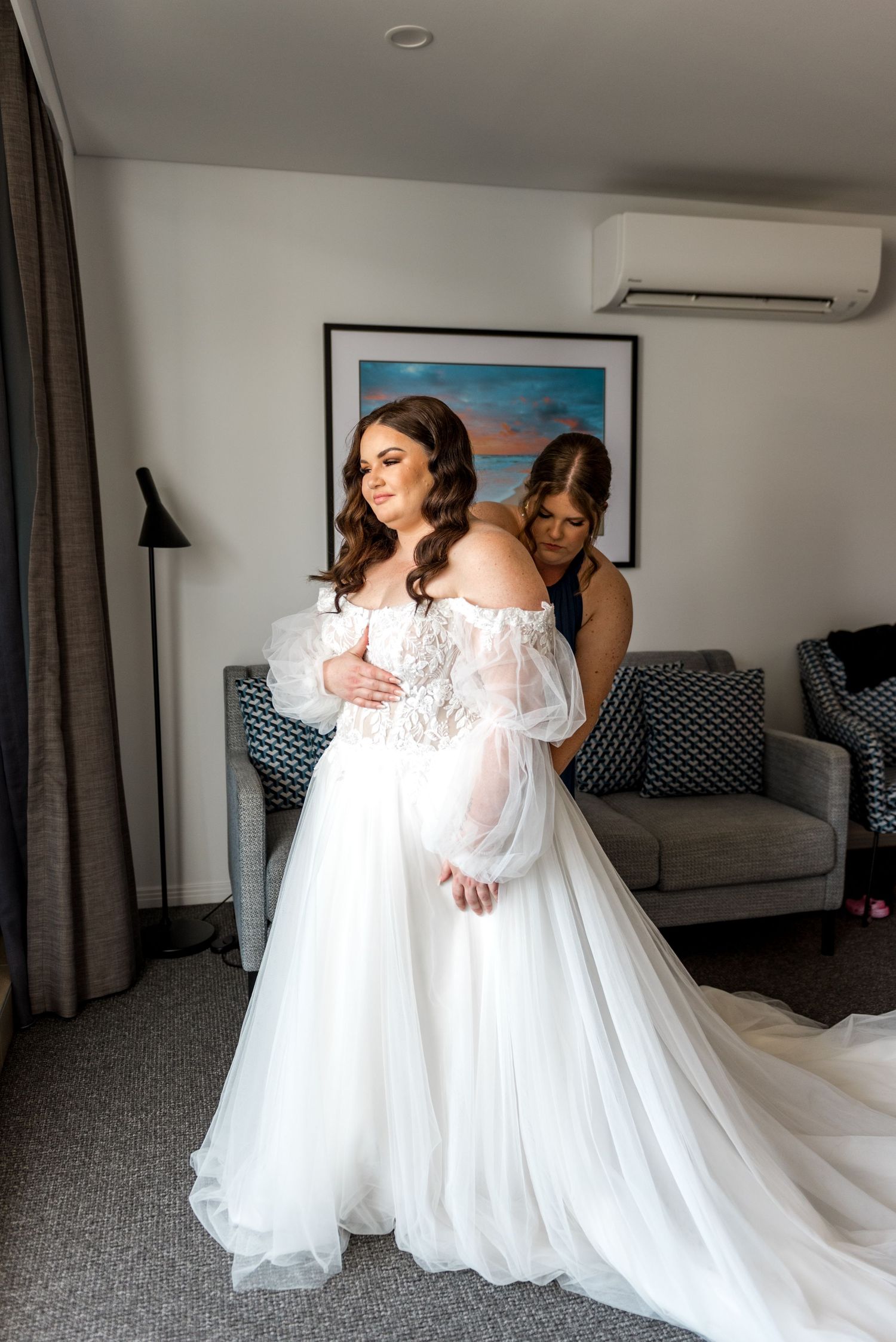 Bride in off-shoulder lace tulle wedding dress being helped by bridesmaid in hotel room.