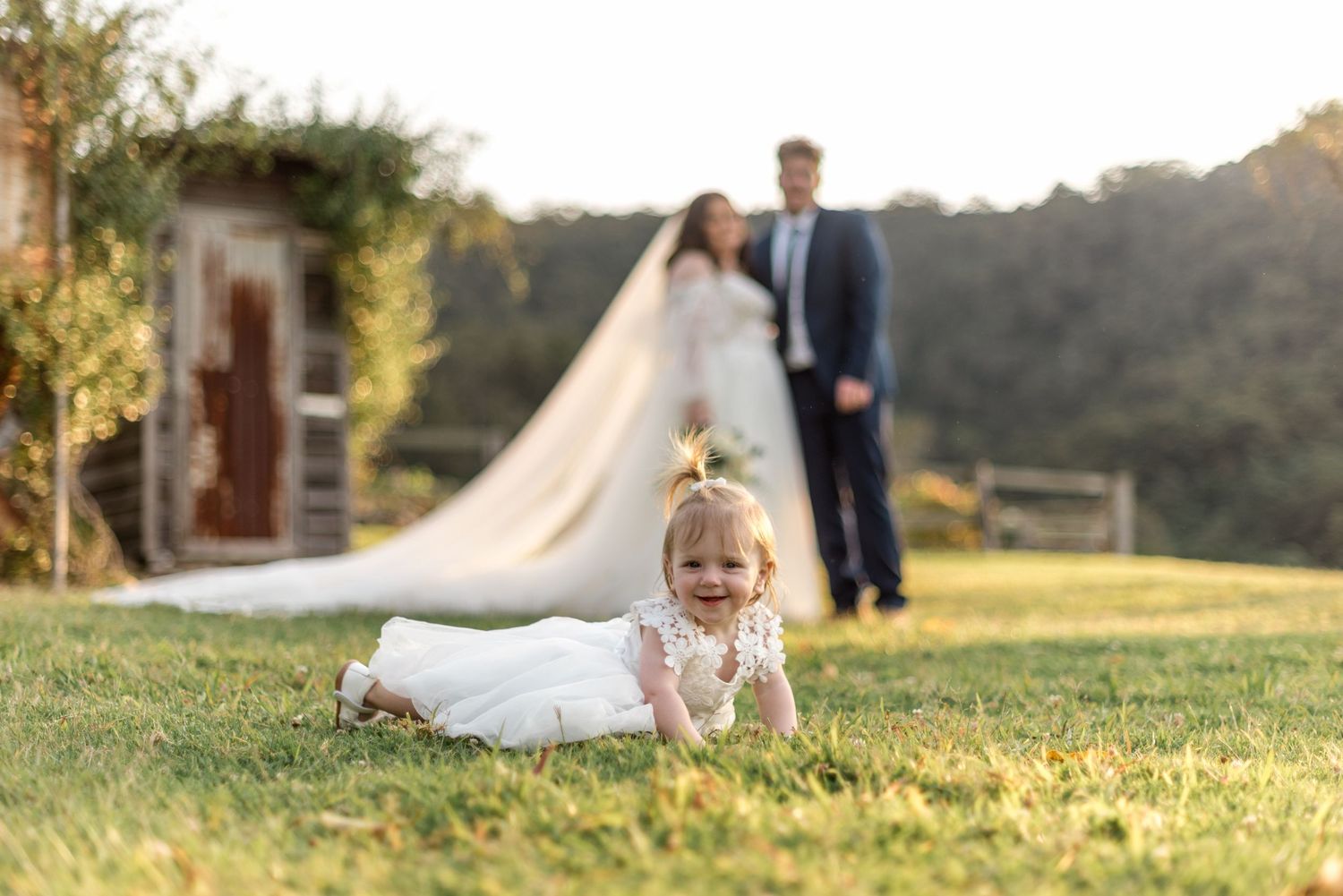 Smiling baby in white dress crawling on grass with bride and groom in background.