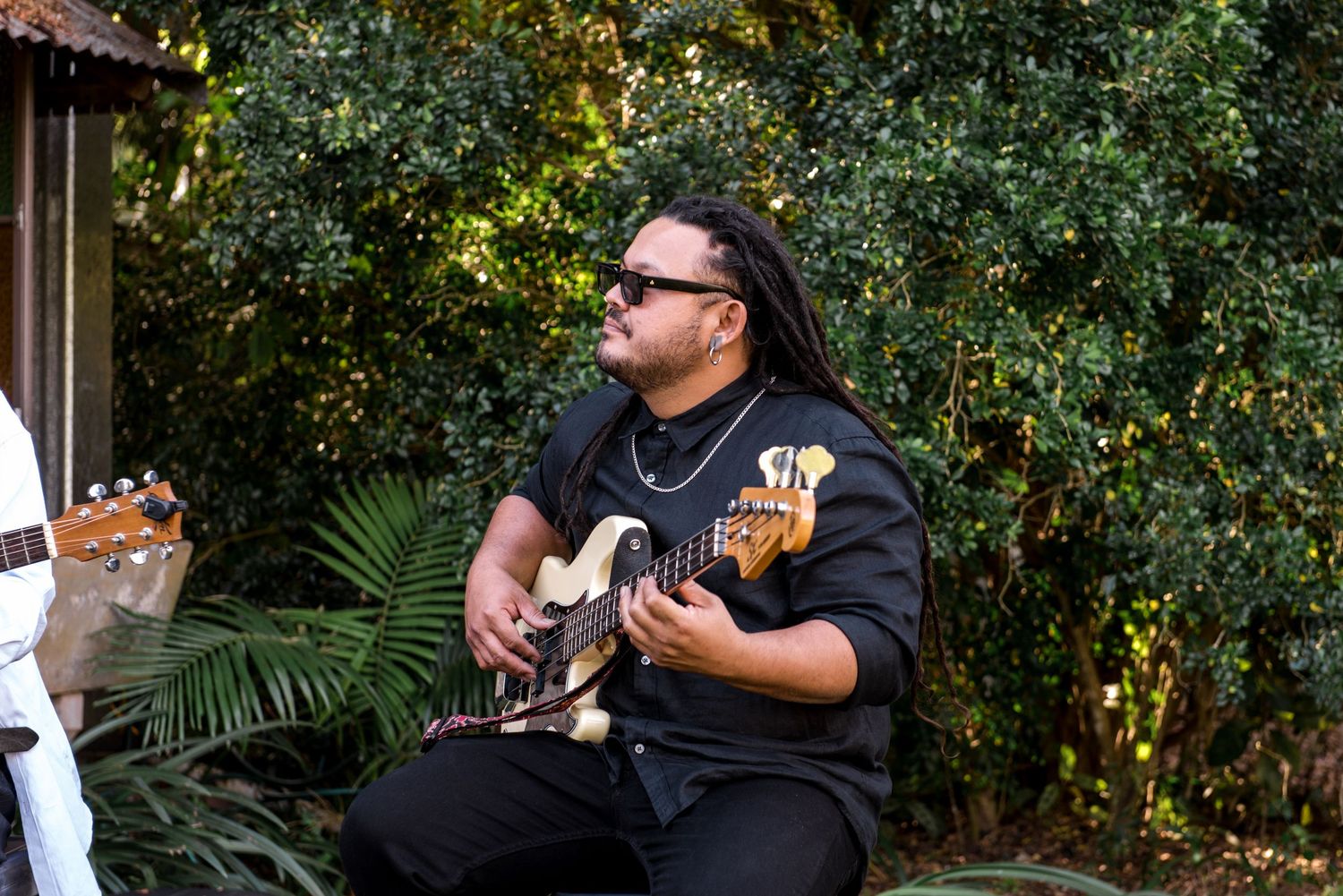 Musician in black plays electric guitar outdoors surrounded by lush green foliage.