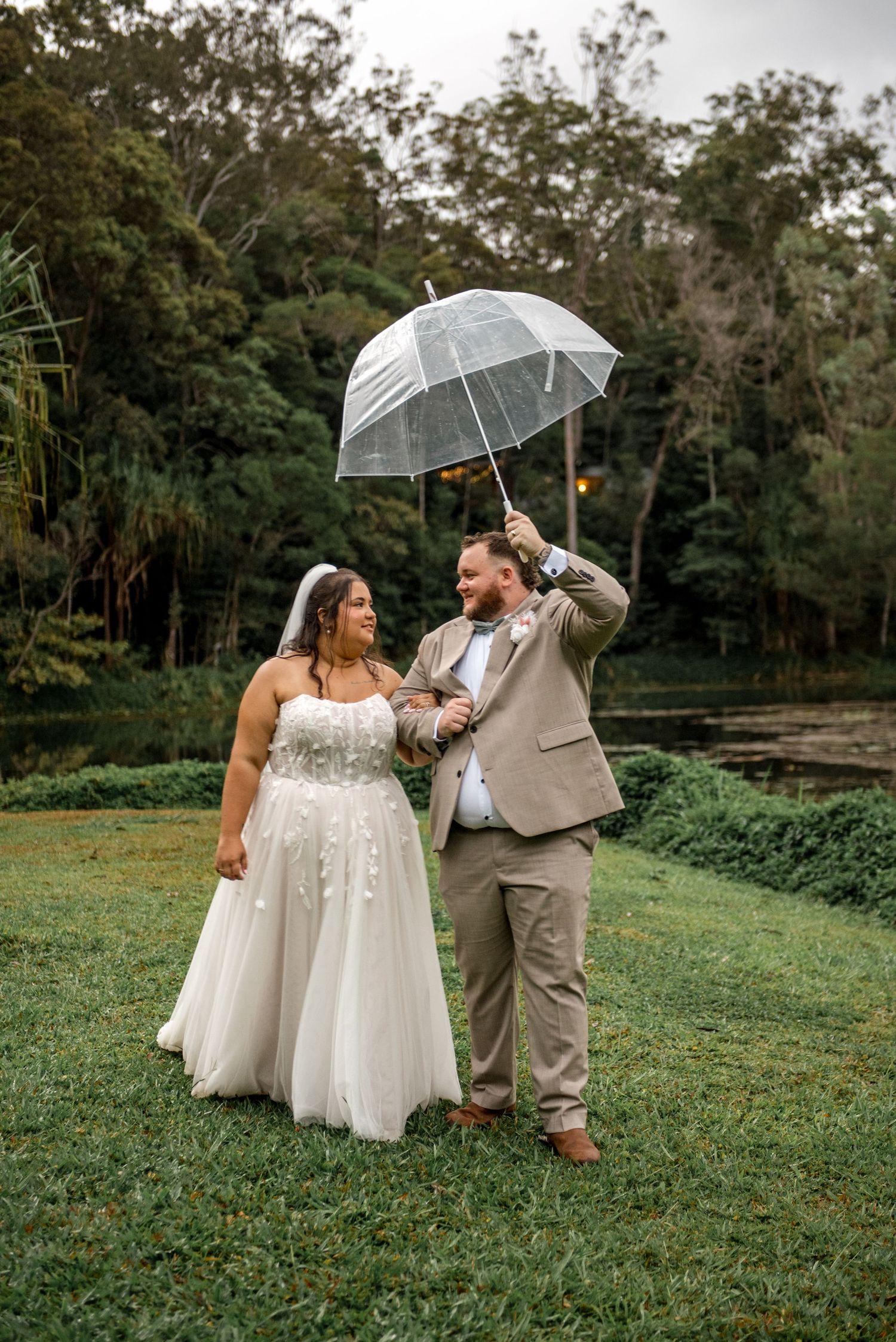Bride and groom share a romantic moment under a clear umbrella on lush green grass with trees in the background.