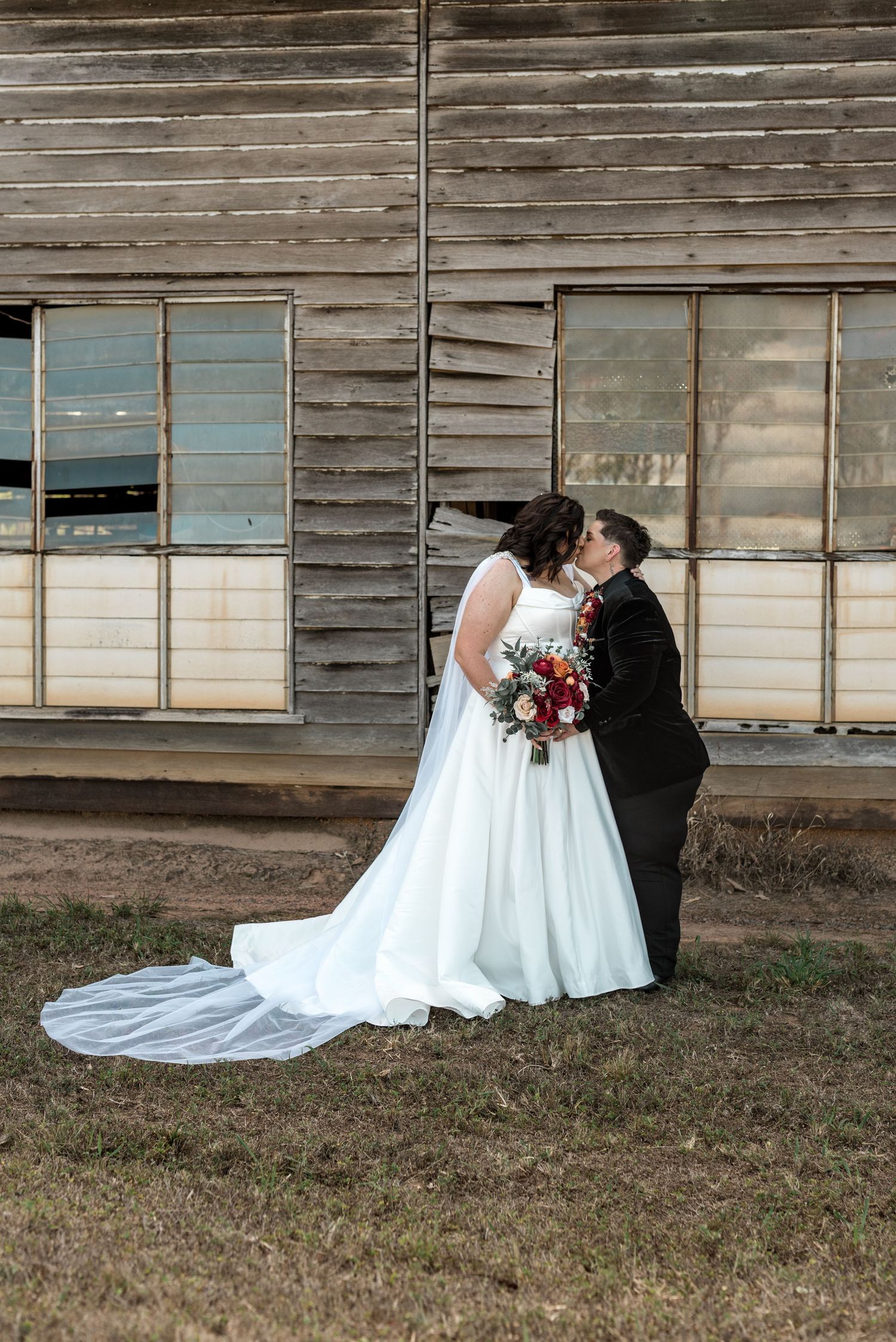 Bride in flowing white gown with long train kisses partner in black suit before a rustic wooden barn wall.