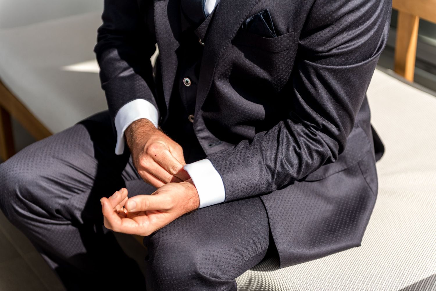 Close-up of a groom in a dark suit adjusting his white shirt cuffs while seated.