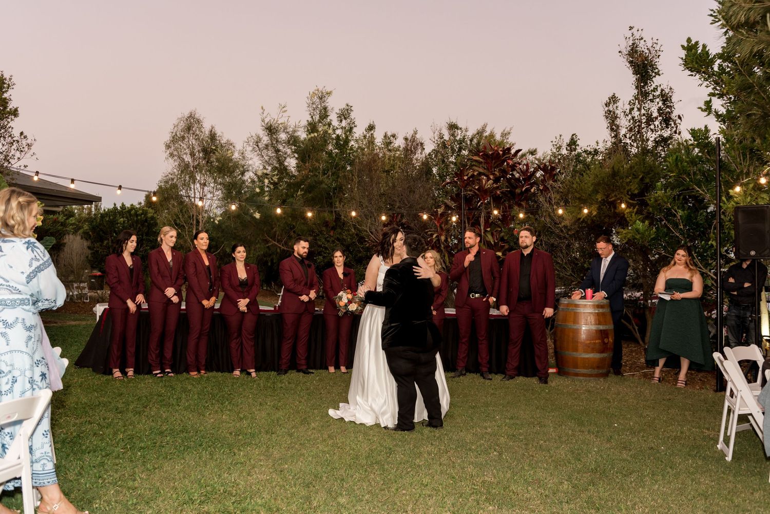 Bride and groom share first dance on lawn with wedding party in burgundy suits watching.