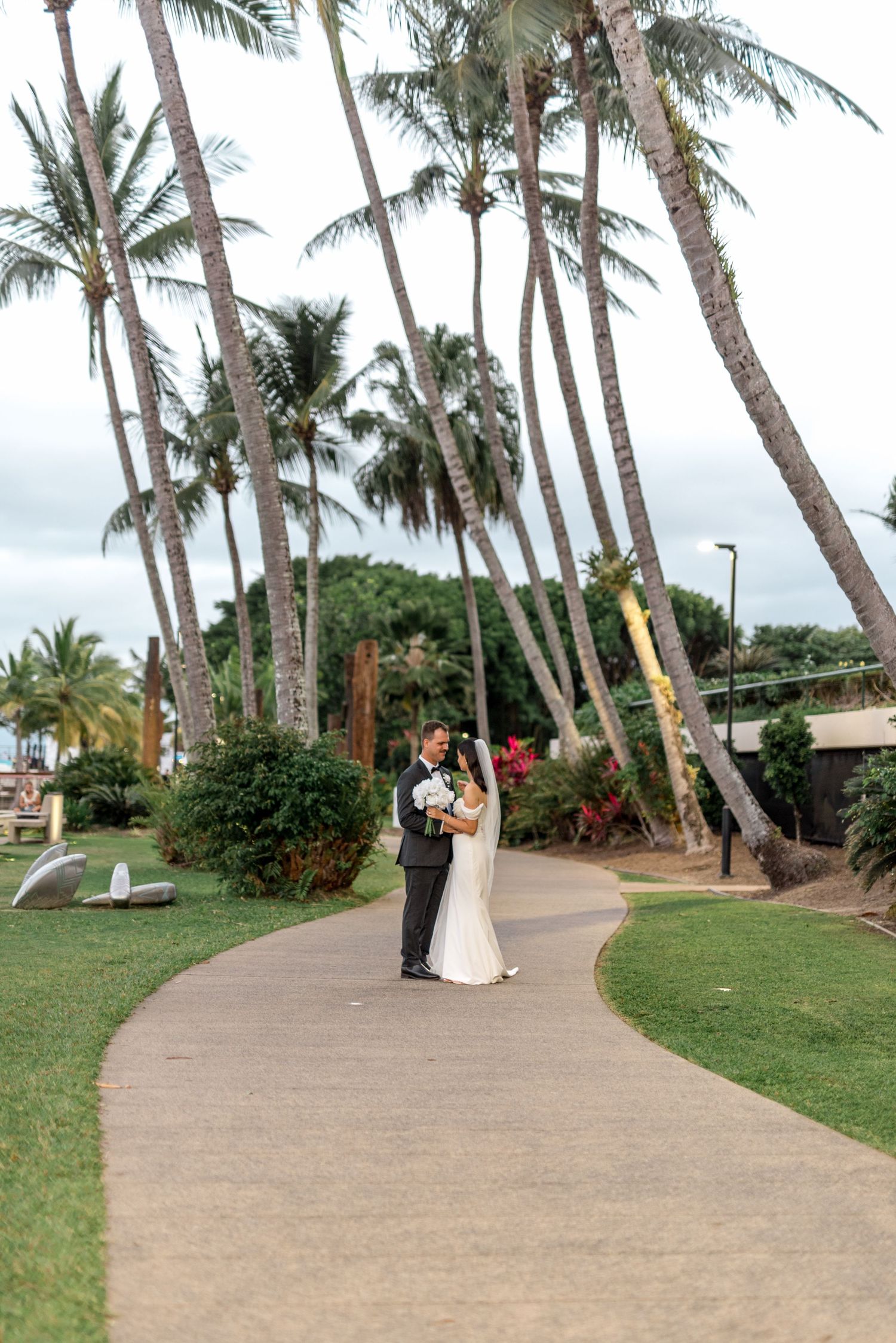 Bride and groom embracing on a winding pathway surrounded by lush tropical palm trees at an outdoor Hawaii wedding.