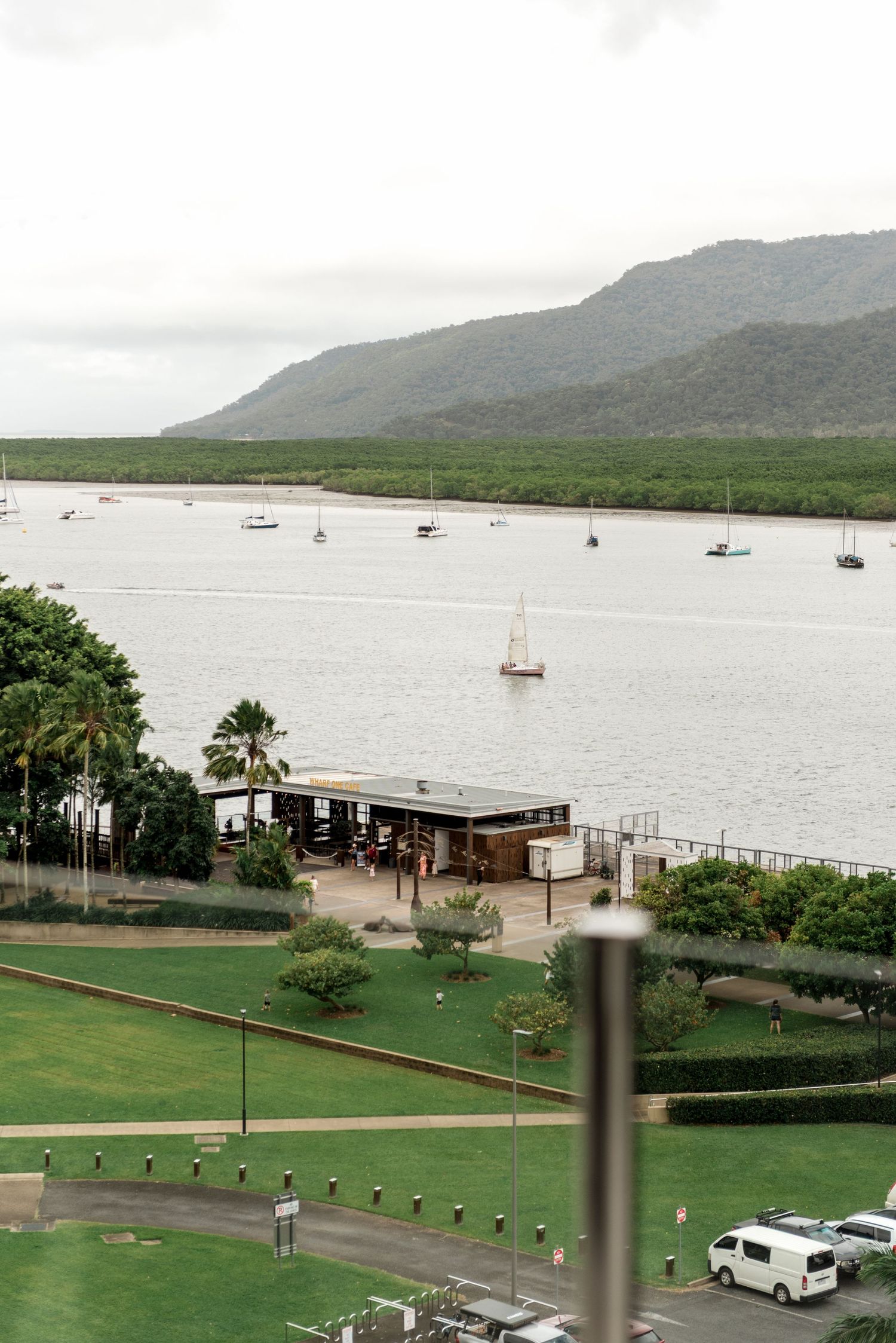 Scenic waterfront view from a high-rise balcony showing sailboats, lush greenery, and misty mountains in the background.