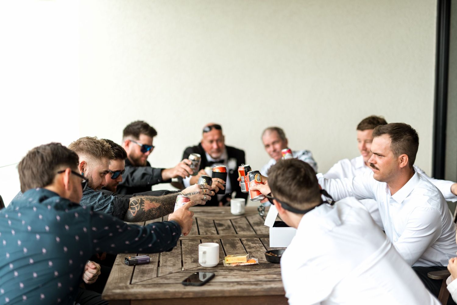 Group of groomsmen toasting drinks around a rustic wooden table on an outdoor terrace.