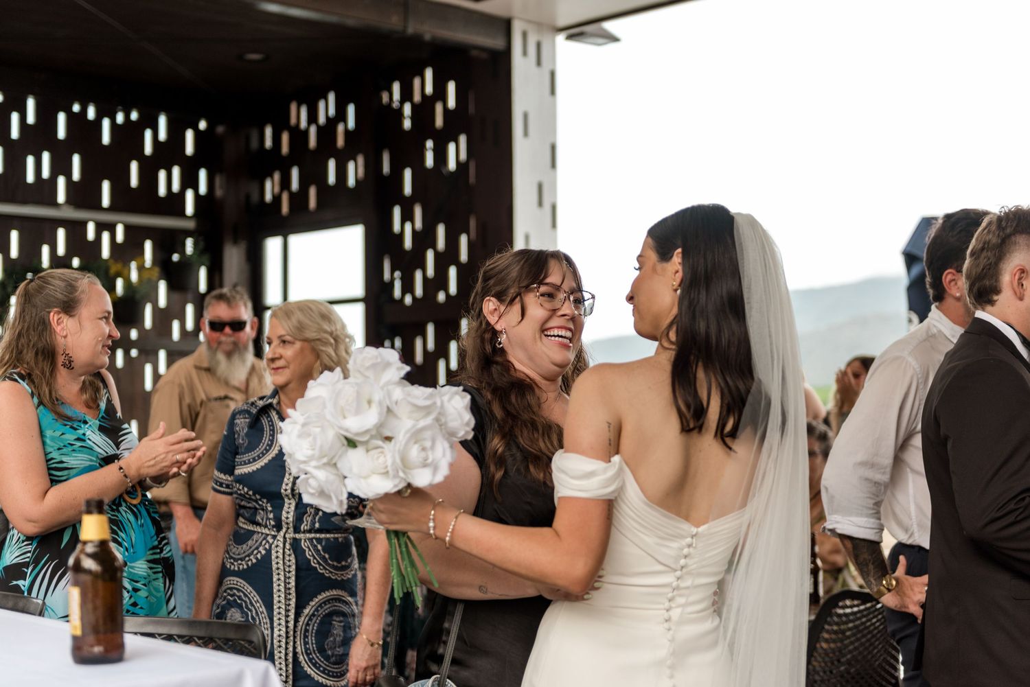 Bride in white off-shoulder gown receiving white bouquet from smiling guest at reception.