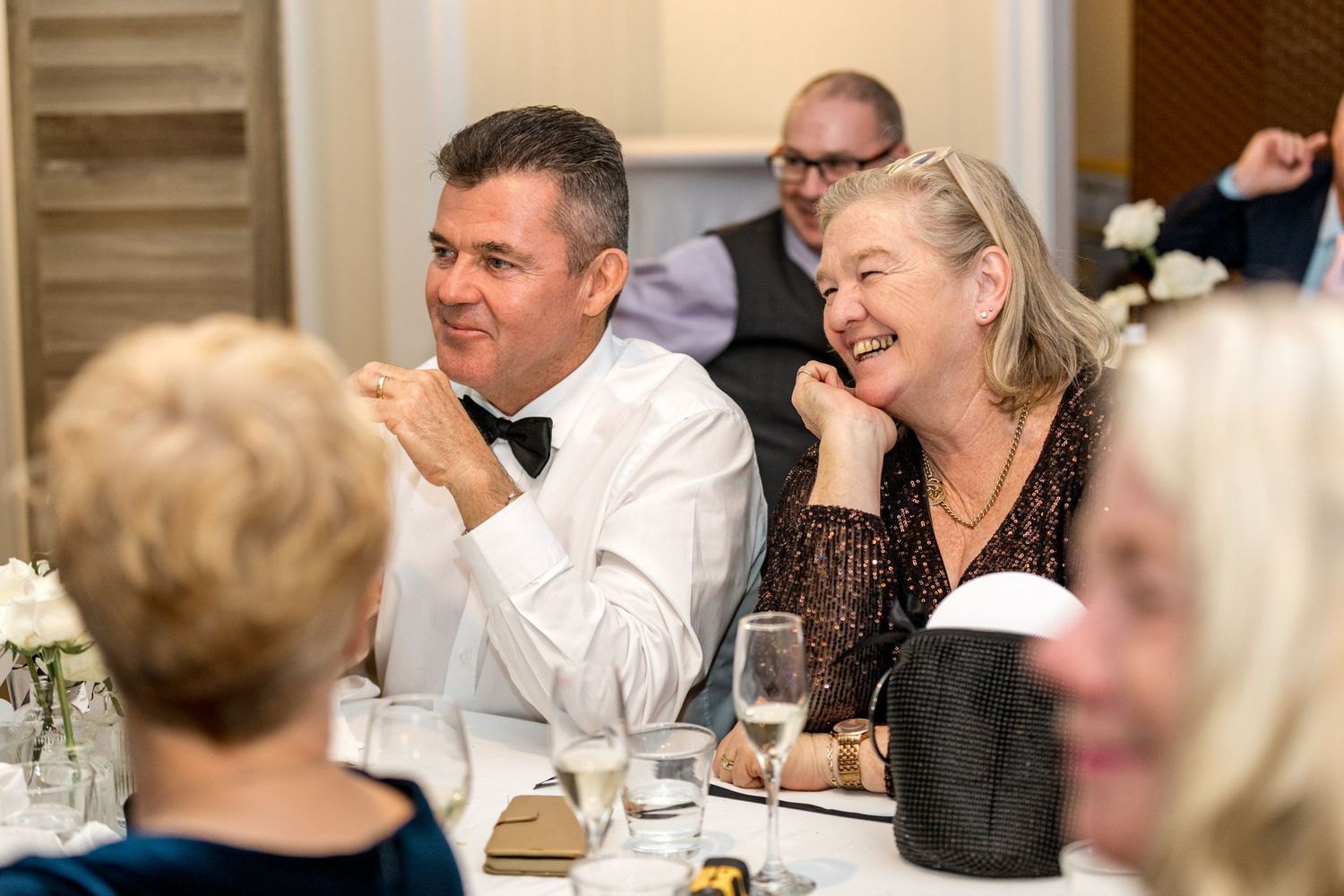 Laughing couple in formal attire enjoying wedding reception dinner with champagne glasses.