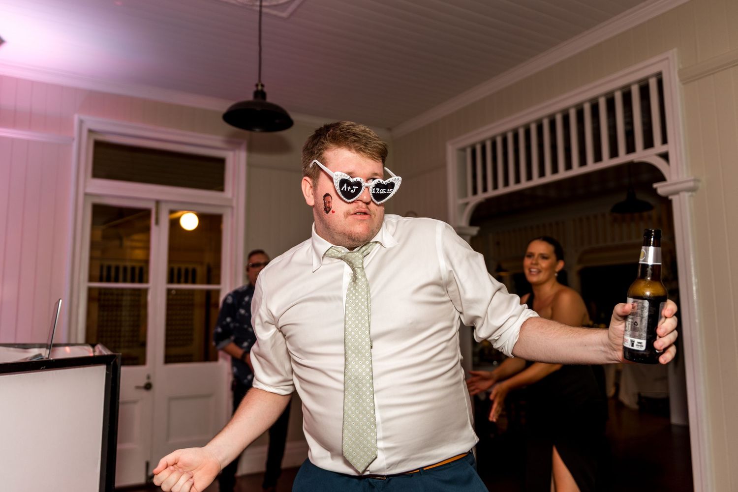 Man dancing at wedding reception wearing heart-shaped sunglasses while holding a beer bottle.