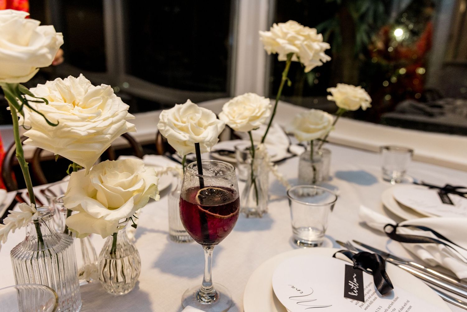 Wedding reception table with white roses, red wine glass, and place settings on white linen.