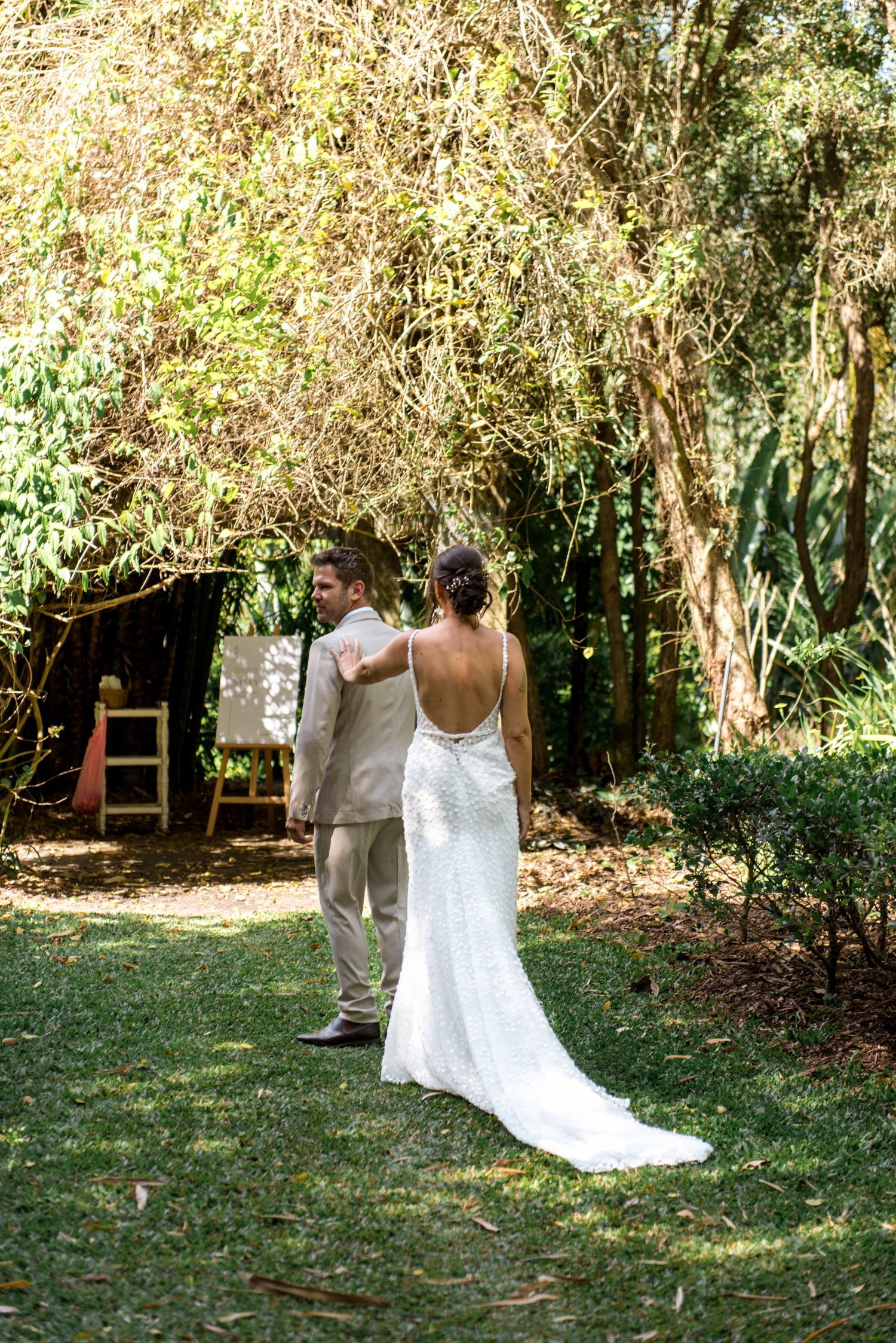 Bride in backless lace gown taps groom's shoulder for first look moment in lush garden setting.