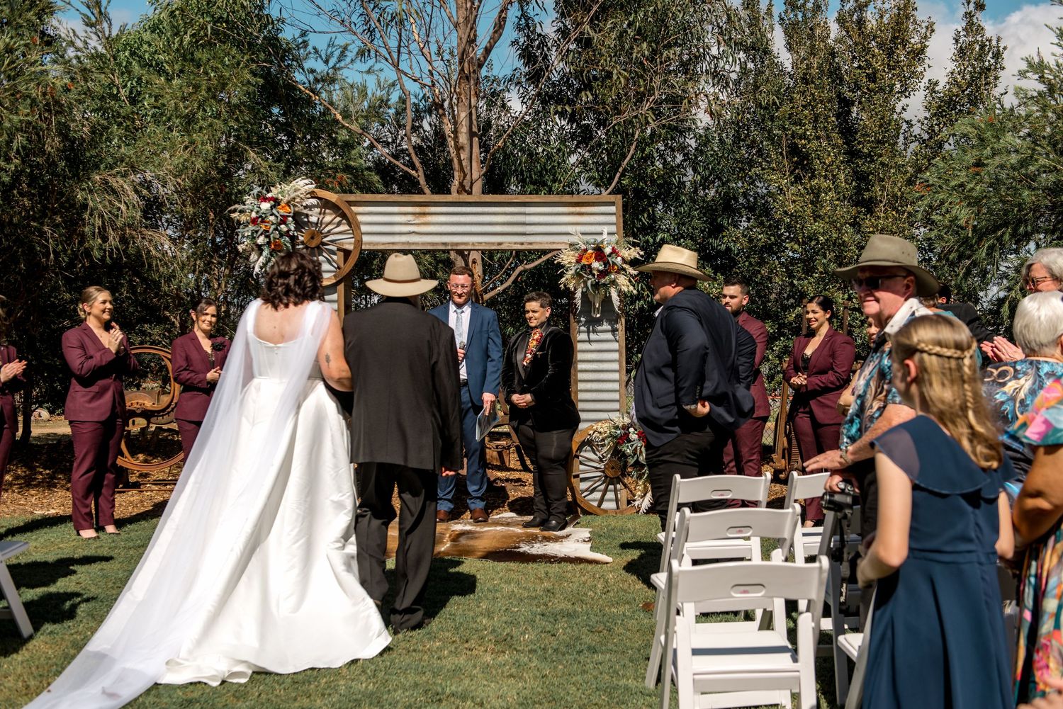 Bride and father walk down outdoor aisle toward groom at rustic corrugated iron wedding altar.