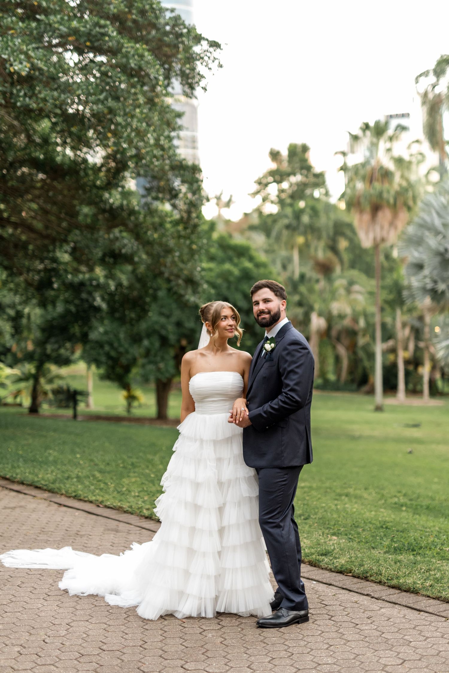 Bride in layered ruffled gown and groom in navy suit pose together in a tropical garden setting.