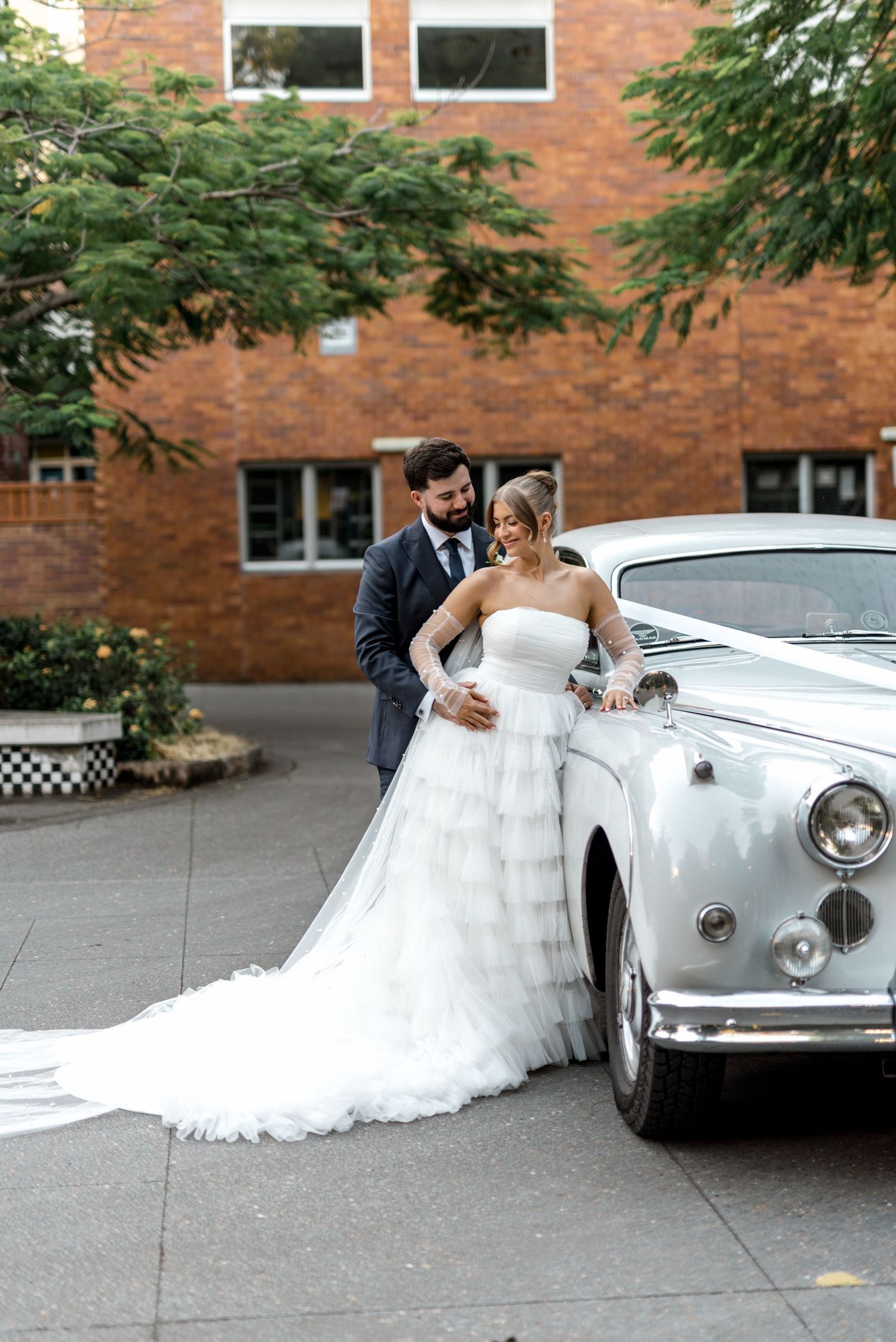 Bride in tiered tulle gown leans against vintage white car with groom in navy suit at outdoor wedding.