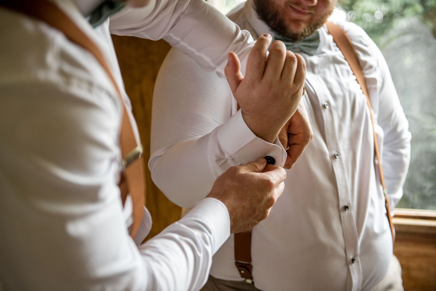 Groomsman fastening cufflinks on groom's white dress shirt sleeve during wedding preparations.
