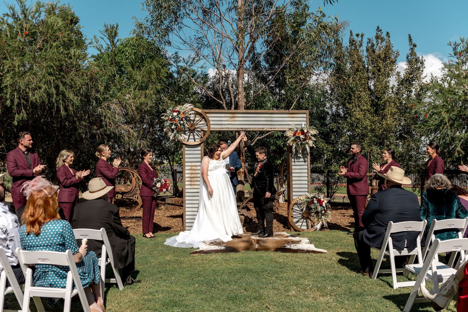 Bride and groom celebrate at outdoor wedding ceremony altar surrounded by bridal party in burgundy attire.