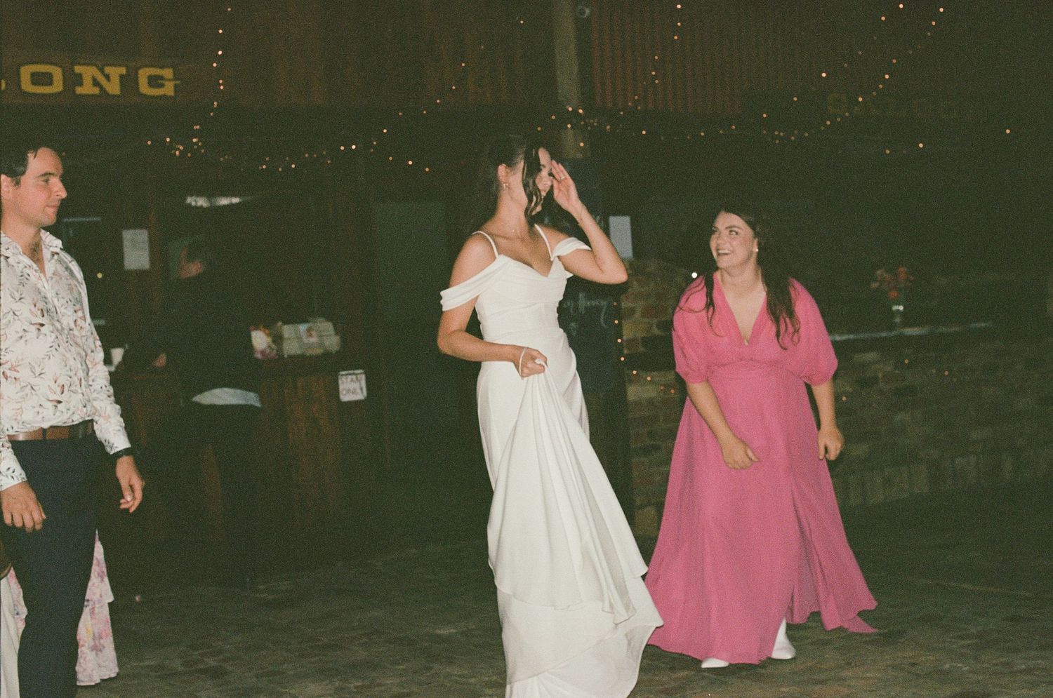 Bride in white gown dancing with guest in pink dress at rustic barn wedding reception.