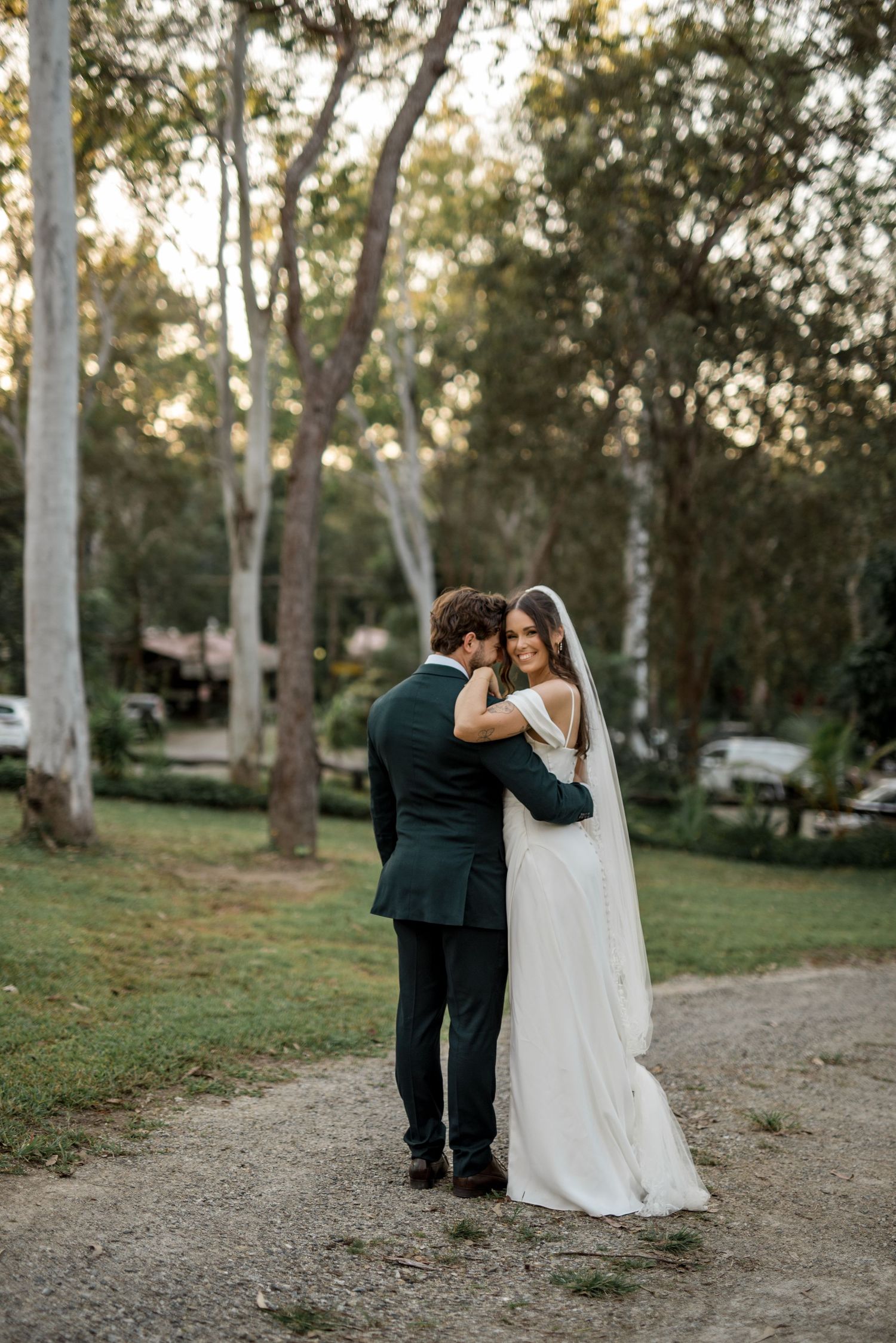 Groom embracing bride from behind as she smiles at camera in a tree-lined garden.