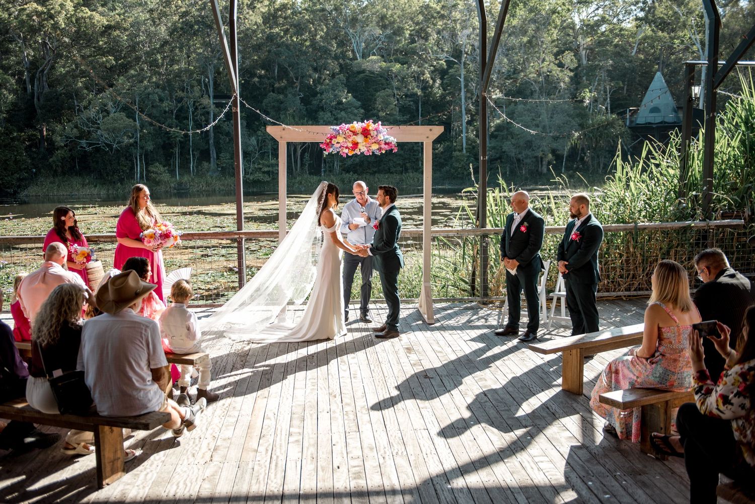 Bride and groom exchange vows at floral arch on wooden deck surrounded by seated wedding guests.