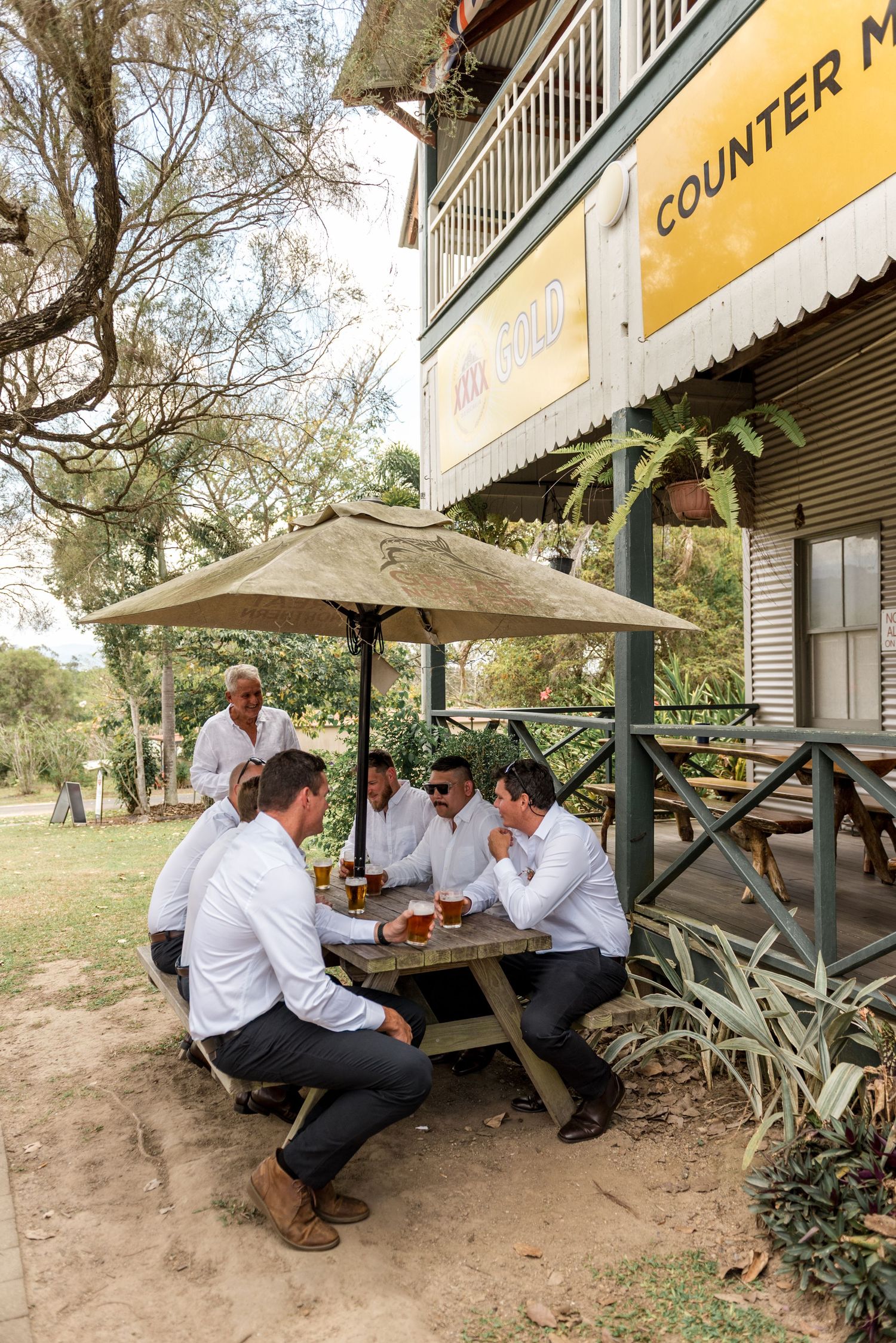 Groomsmen in white shirts enjoying beers at an outdoor picnic table under an umbrella at a country pub.