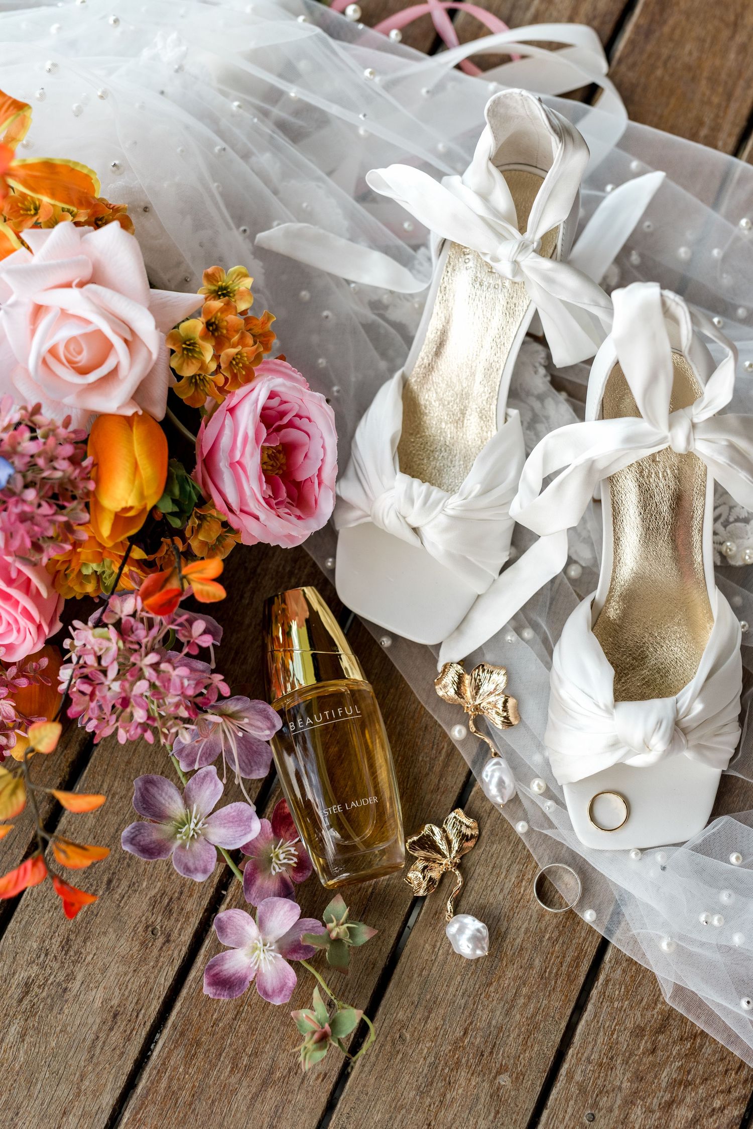 Wedding detail flat lay with white bow heels, veil, Beautiful perfume, gold earrings, and colorful flowers.