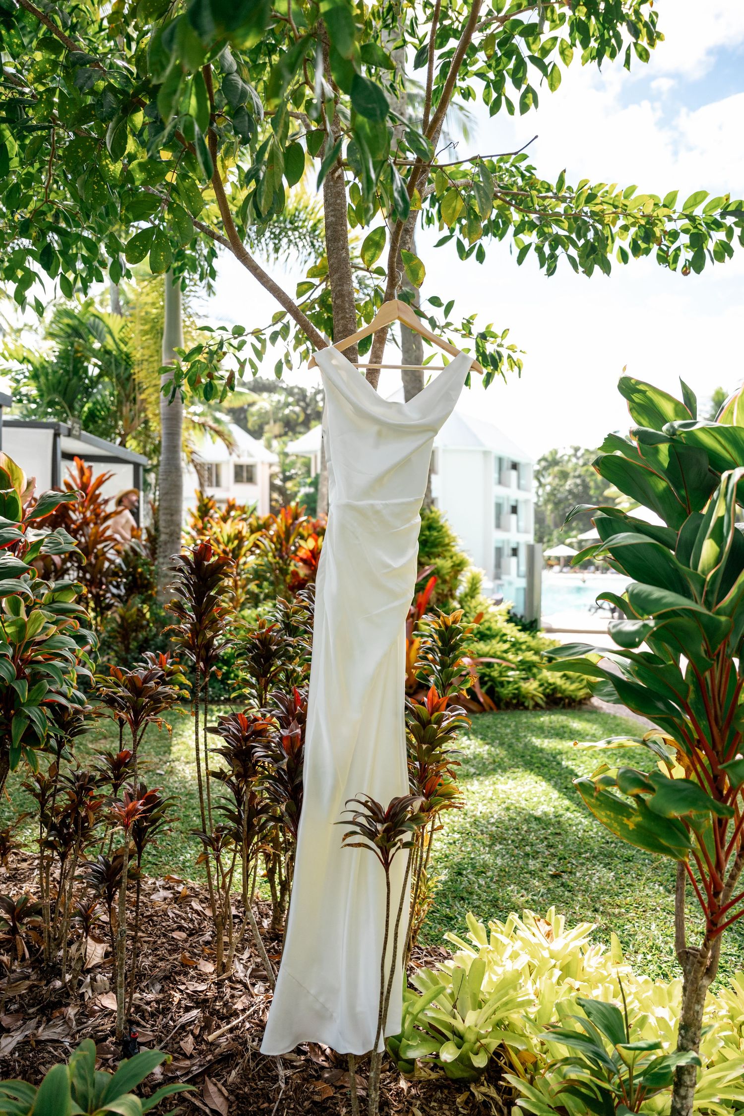 Sleek white bridal dress on wooden hanger displayed outdoors in lush tropical garden with colorful foliage.