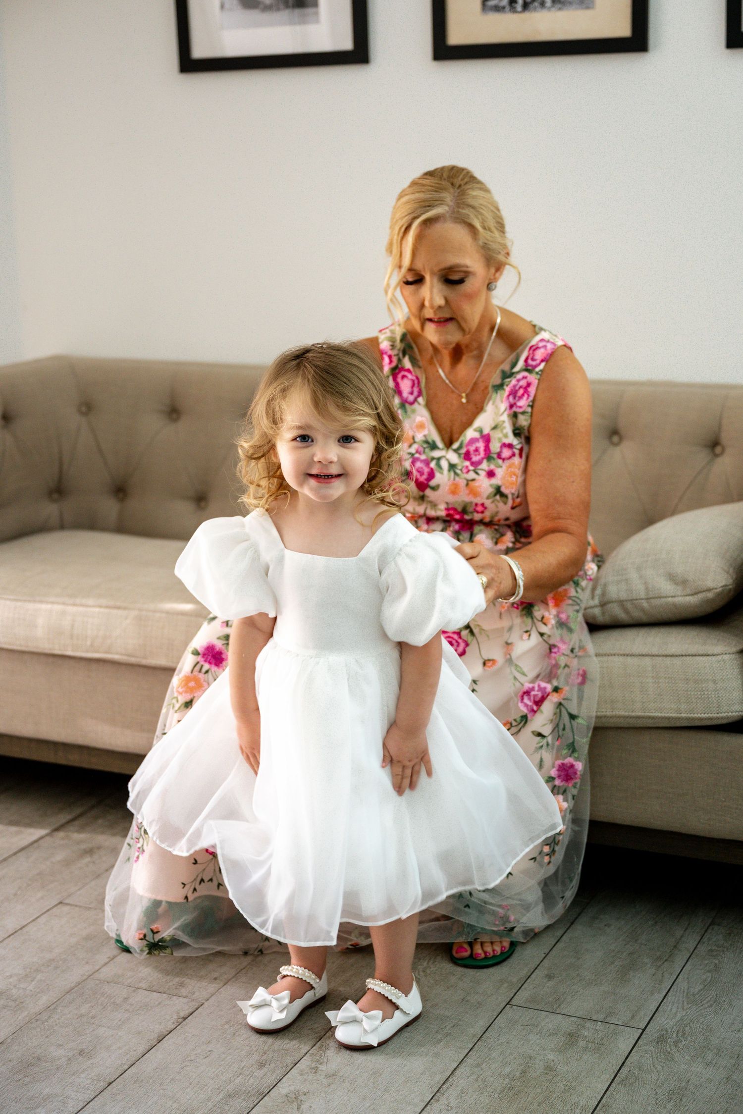 Adorable toddler flower girl in a white dress standing in front of a seated blonde woman in floral dress.