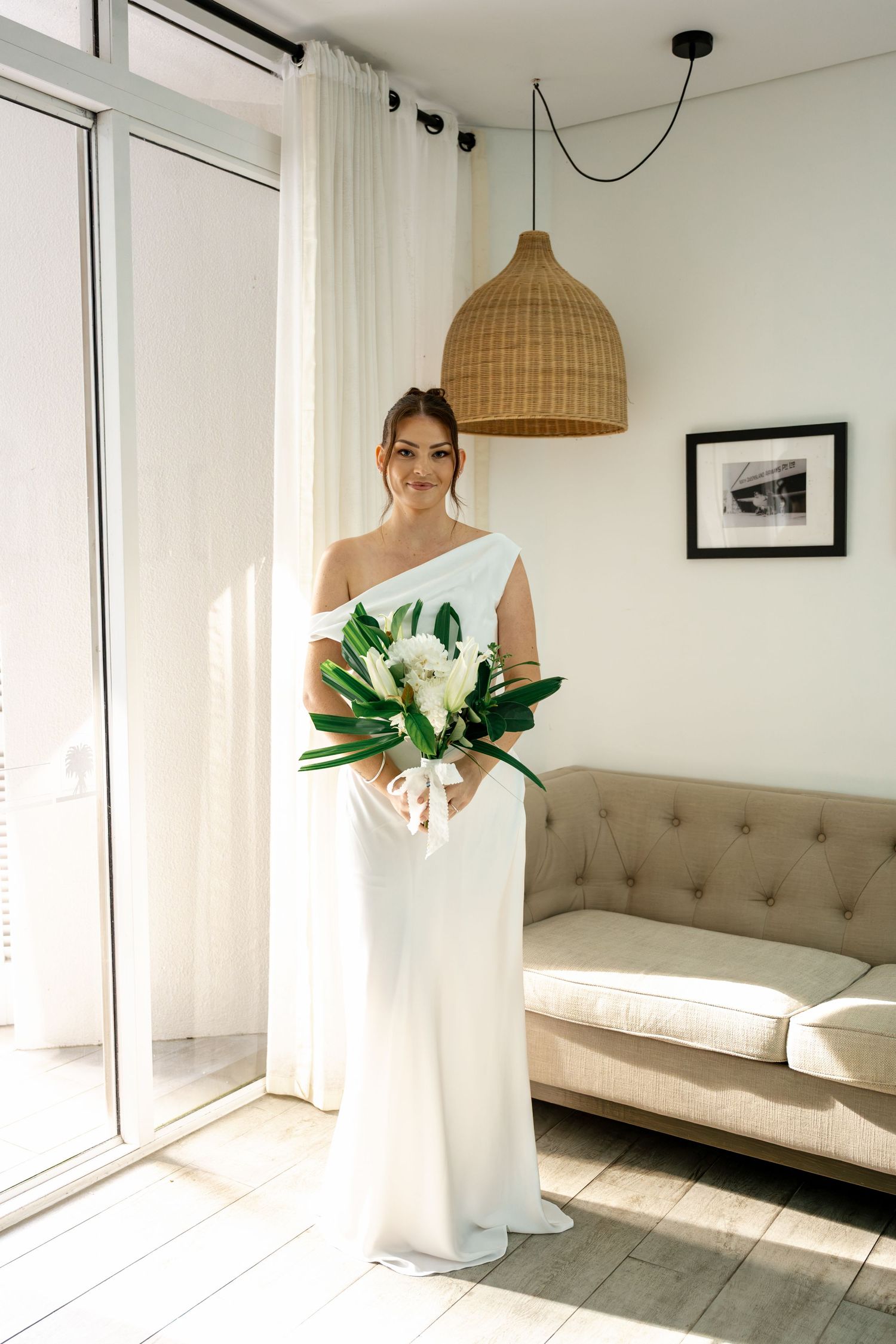 Beautiful bride in minimalist white asymmetric wedding dress holding tropical bouquet standing by bright window at Sheraton Mirage Resort.